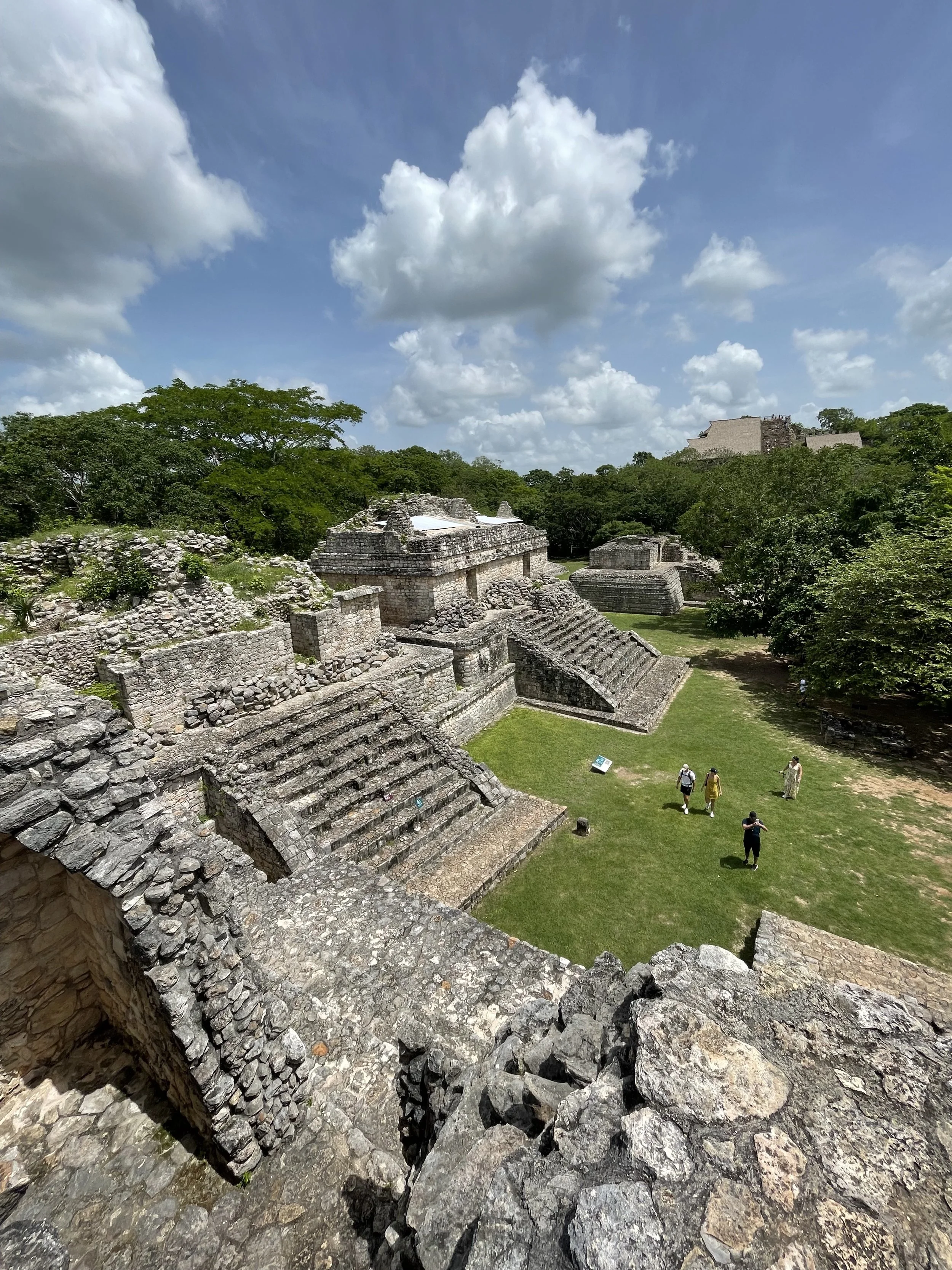 Ancient Mayan ruins with stone stepped pyramids and structures in a lush green landscape under a blue sky with scattered clouds, with a few tourists exploring.