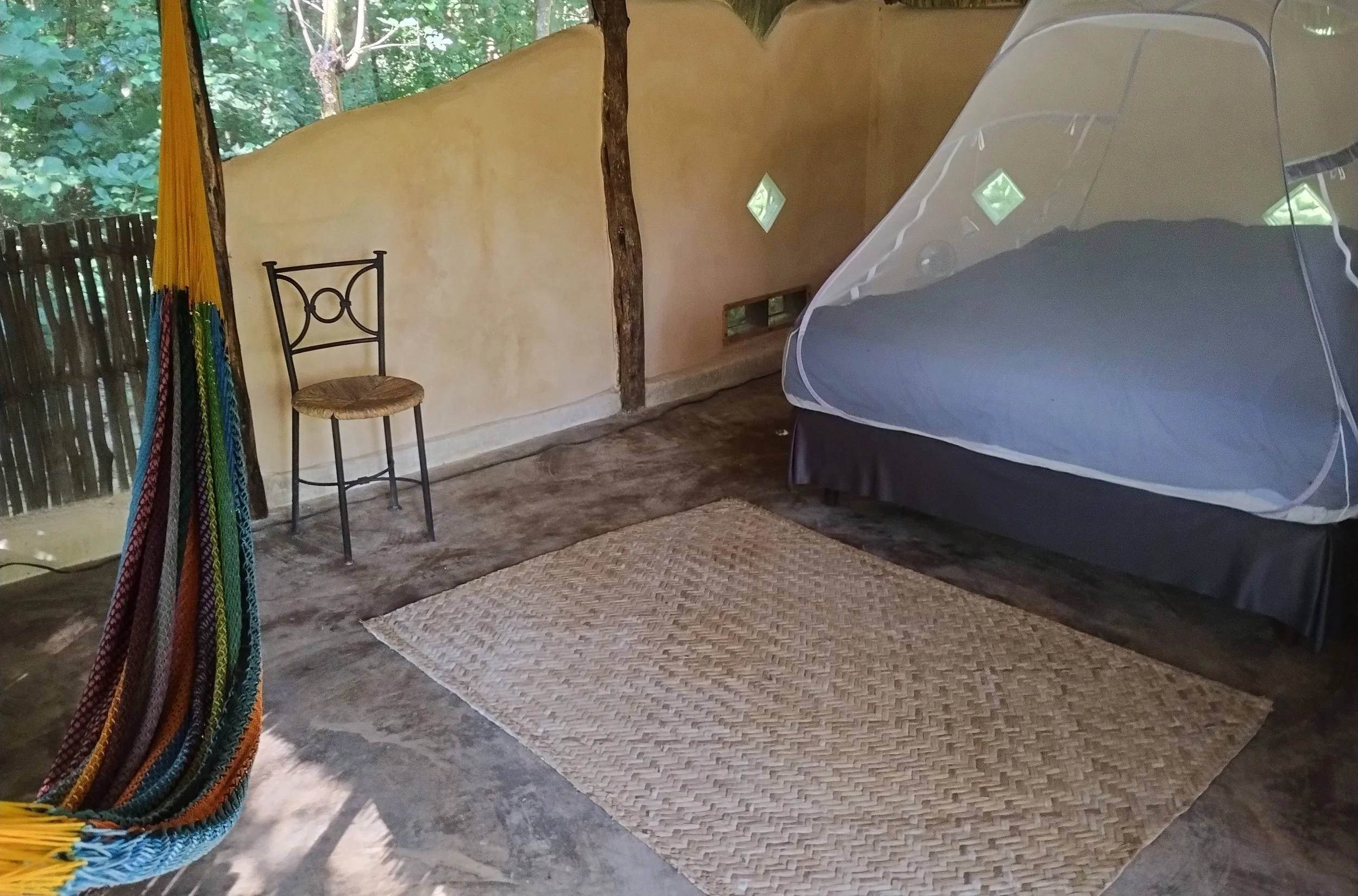 A hammock hangs in front of a bed within a rustic, thatched-roof hut with circular windows, with a small nightstand holding a TV on the right side.