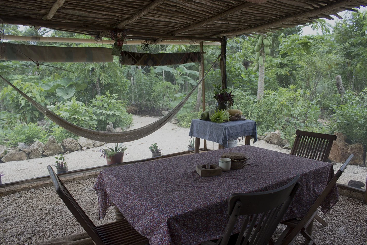 A rustic outdoor dining area with a table covered in a floral tablecloth, surrounded by wooden chairs, overlooking a lush green garden. There is a hammock hanging from the ceiling and an outdoor table with plants and gourds in the background.