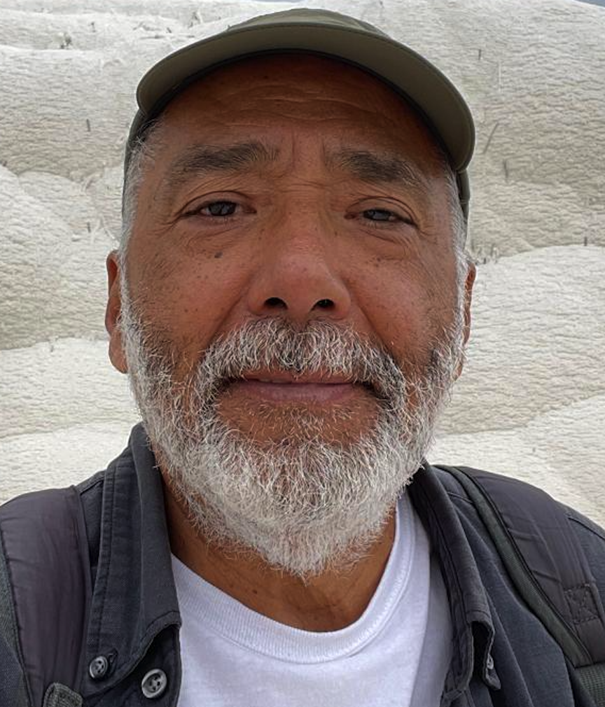 Close-up of an elderly Asian man with gray beard and mustache, wearing a beige cap and gray backpack, smiling in front of a white wall.