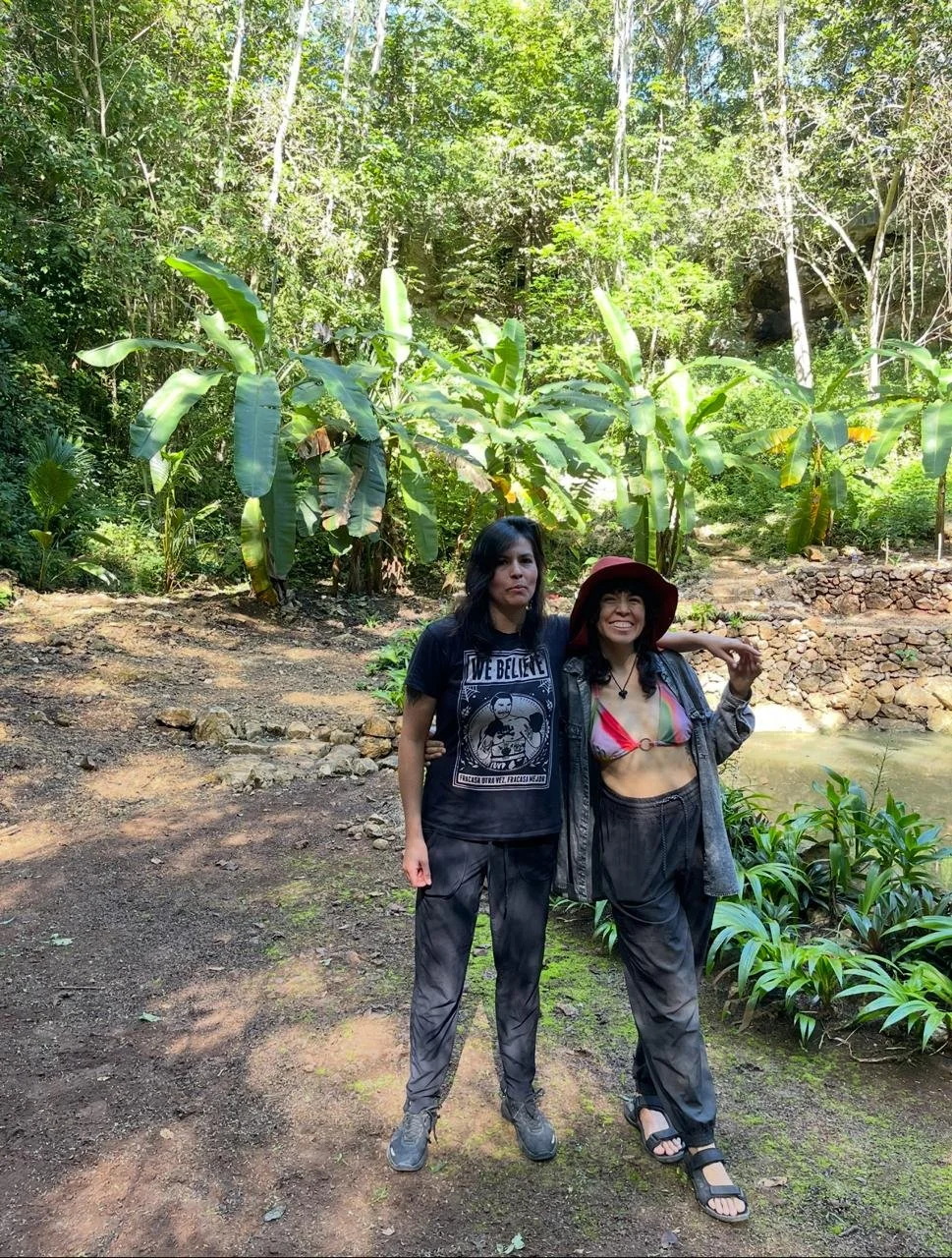 Two women standing together on a dirt path in a lush, green forest with large banana leaves and other foliage, smiling and enjoying the outdoors.