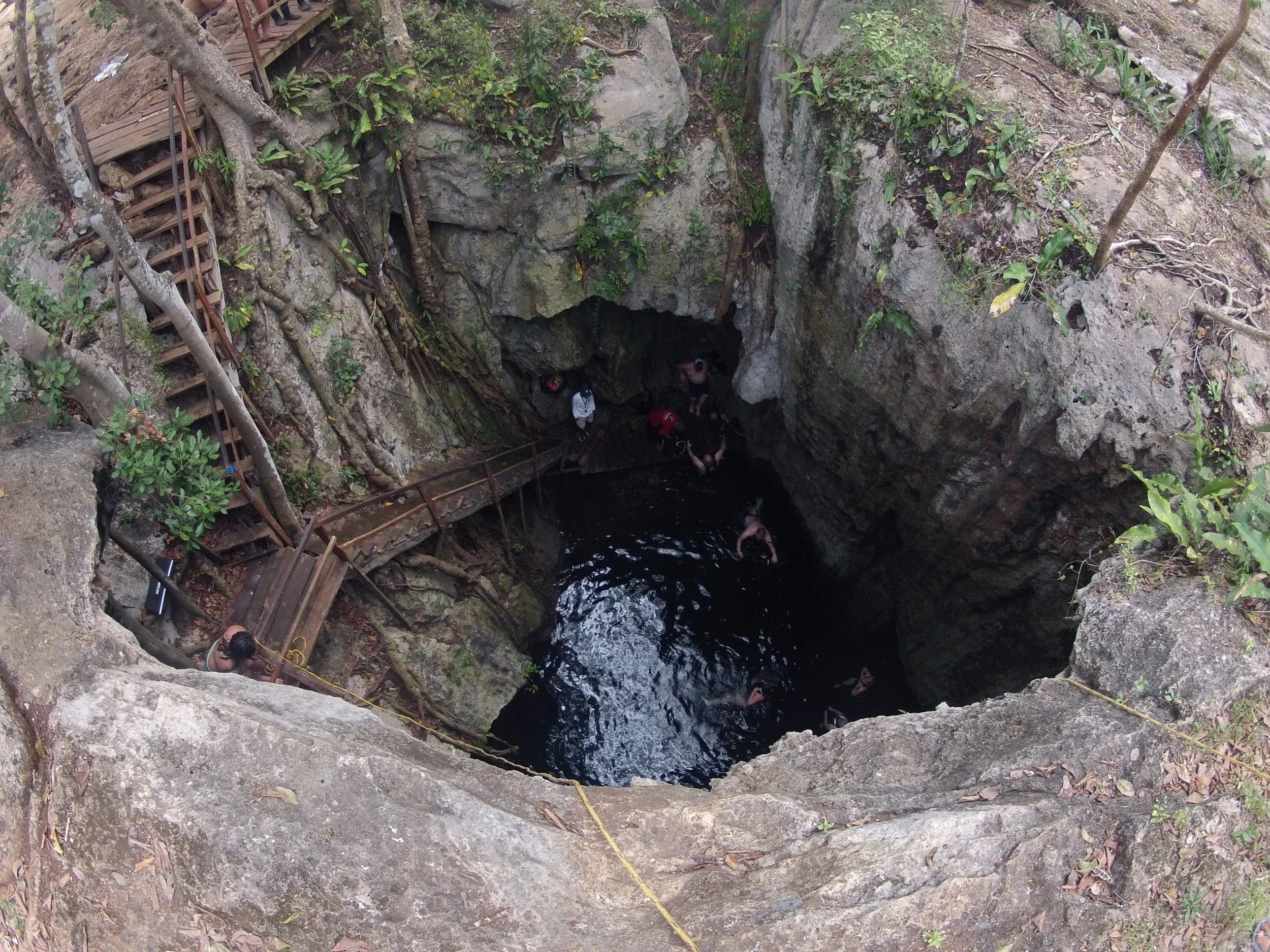People exploring a deep sinkhole with wooden stairs and walkways, surrounded by rocks and greenery.