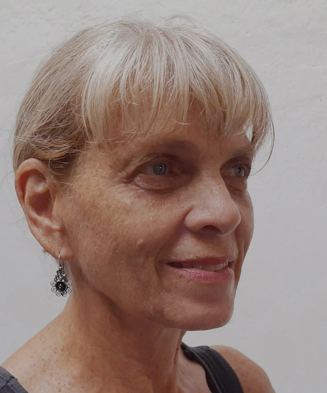 Close-up of a woman with short blonde hair, earrings, freckles, and a black top, smiling slightly against a plain white wall.