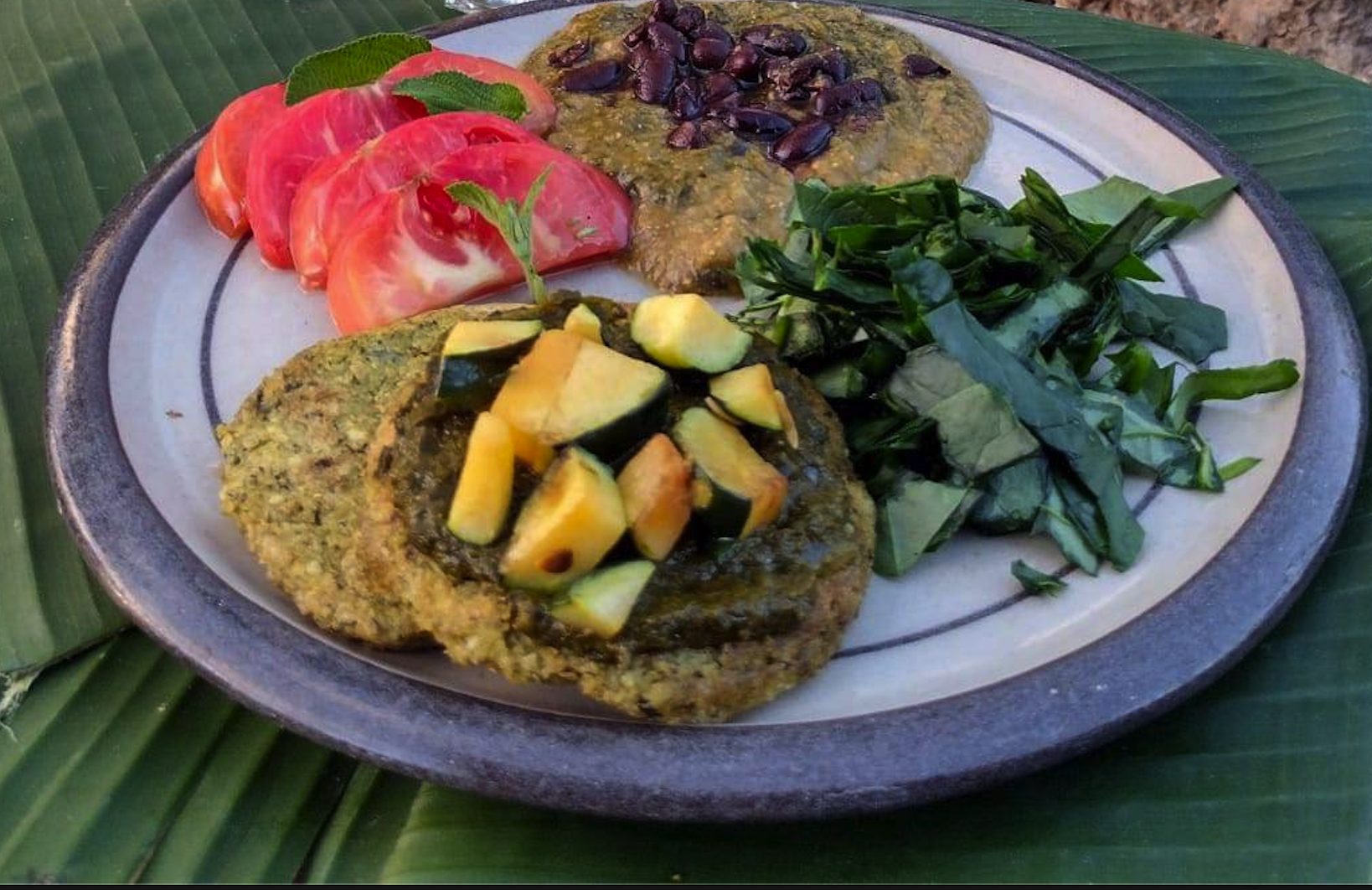 A traditional Latin American dish featuring two breaded and fried pieces topped with diced squash, served alongside sliced tomatoes, chopped greens, and a bean mixture, all arranged on a ceramic platter with a leaf beneath.