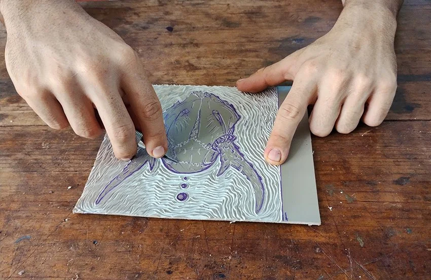 Two hands with light skin are pressing down on a sheet of metal with a scratched and textured surface, on a wooden table. The person is creating an engraved image of a cat's face with a tool.