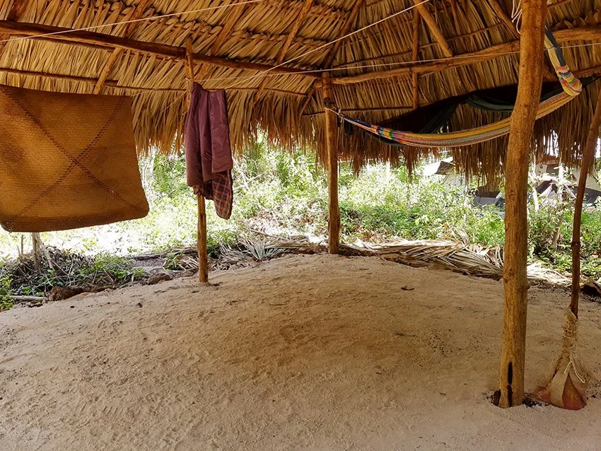A rustic outdoor shelter with a thatched roof made of palm leaves, supported by wooden poles, with clothes hanging and a hammock inside, surrounded by greenery.