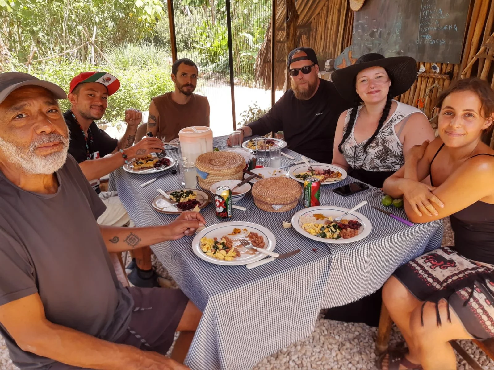 A group of seven people sitting around a dining table outdoors, enjoying a meal, with plates of food, drinks, and a central basket on the table.