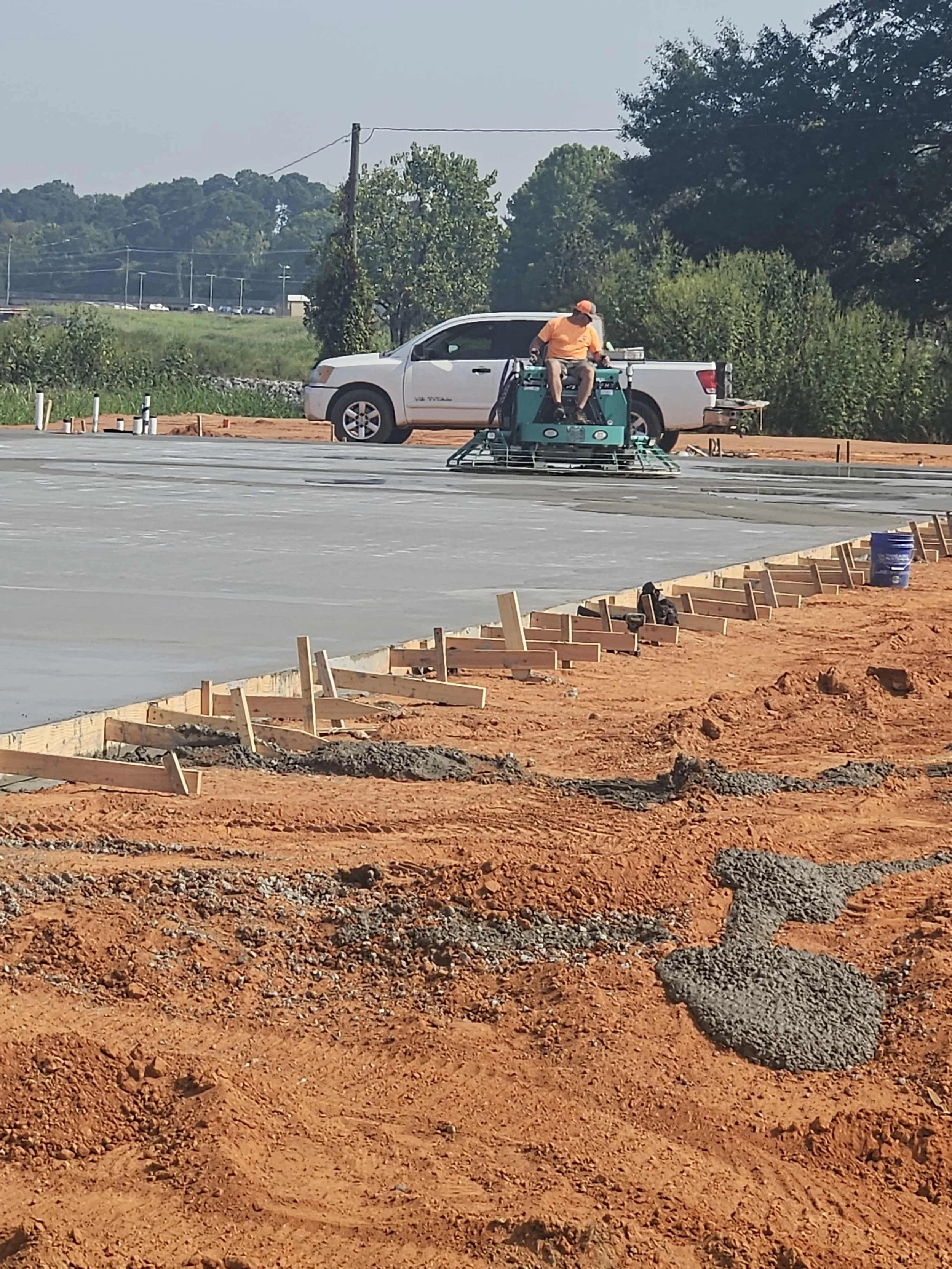 A construction worker operating a ground smoothing machine on a paved surface at a construction site, with a white pickup truck parked behind him, and wooden forms and construction materials on the red dirt ground.