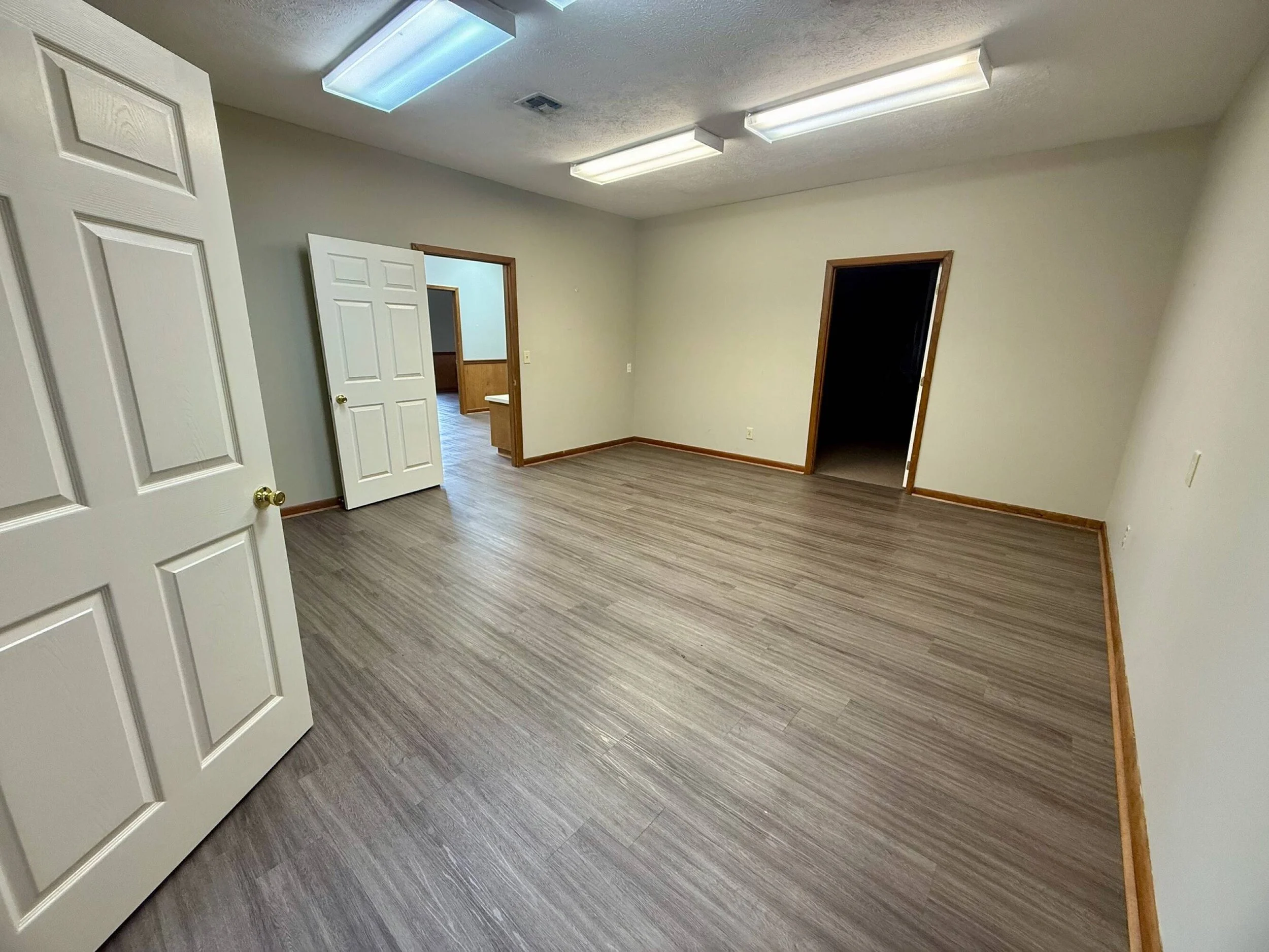 Empty room with wood flooring, white walls, and open doors leading to other rooms, illuminated by fluorescent ceiling lights.