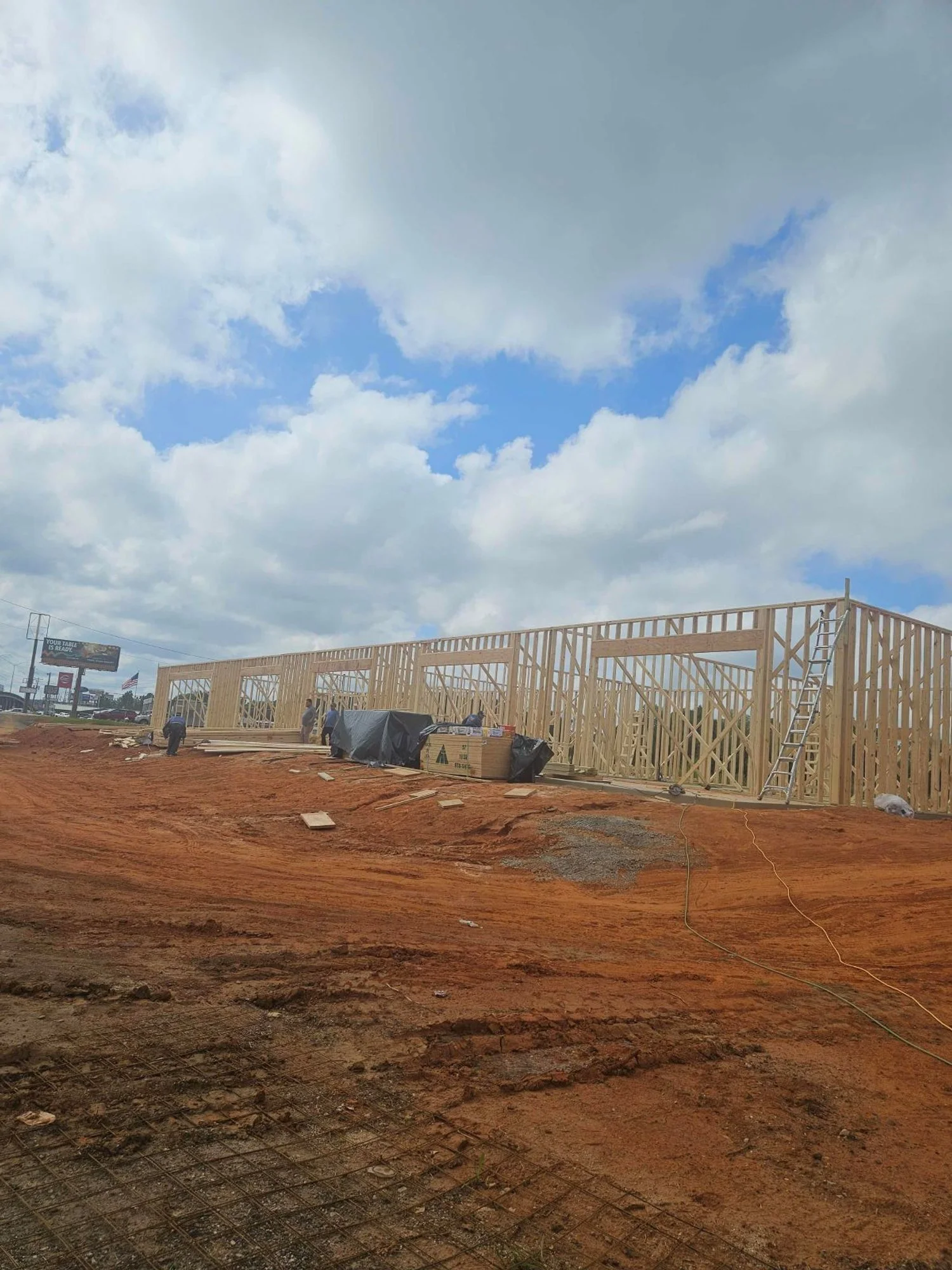 Construction site with a wooden framework for a building, a ladder, and workers working on the structure. The ground is reddish-brown dirt with some materials and equipment around.