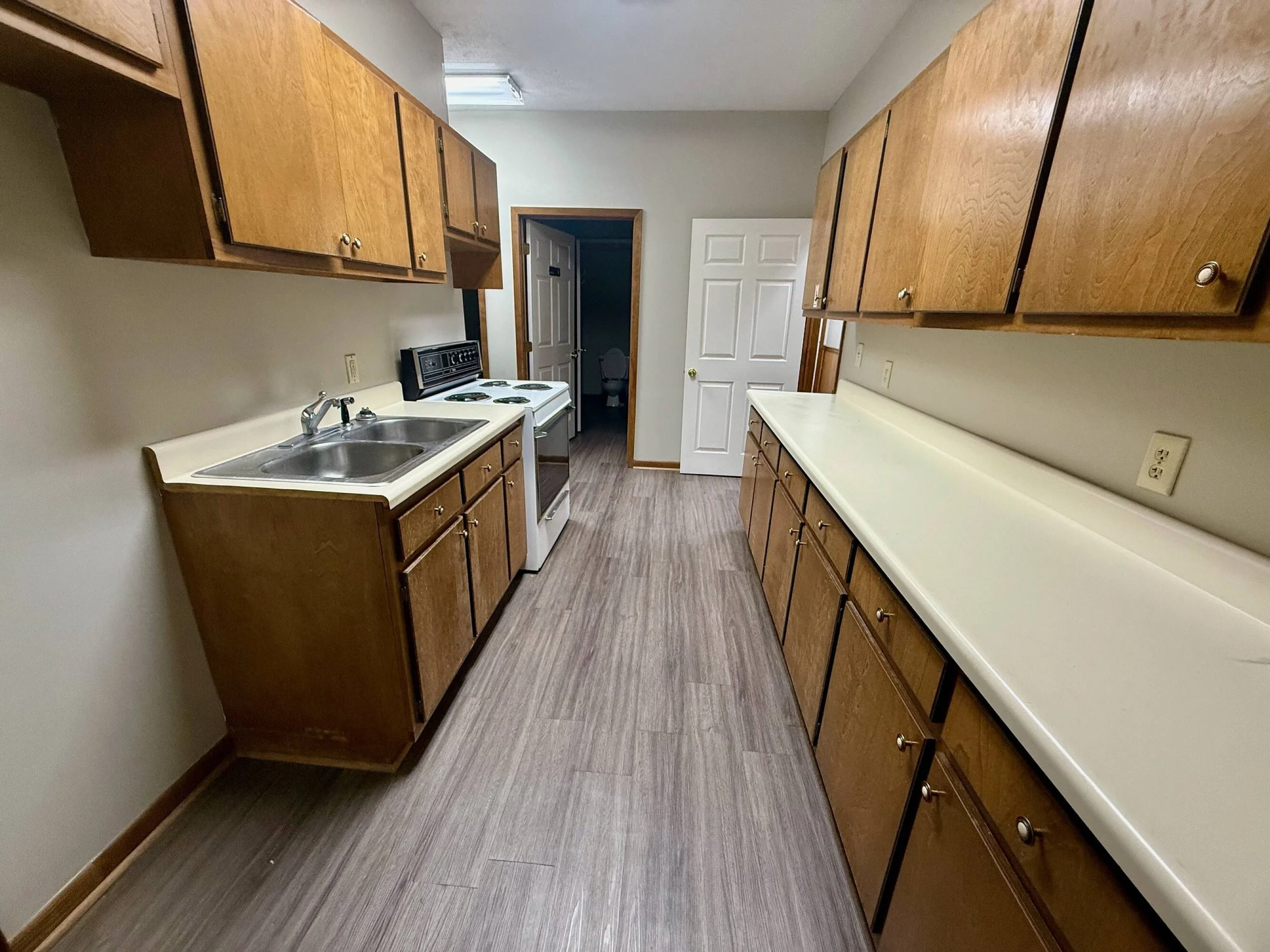 Kitchen with wooden cabinets, white countertops, a sink, an electric stove, and a bathroom visible through an open door.