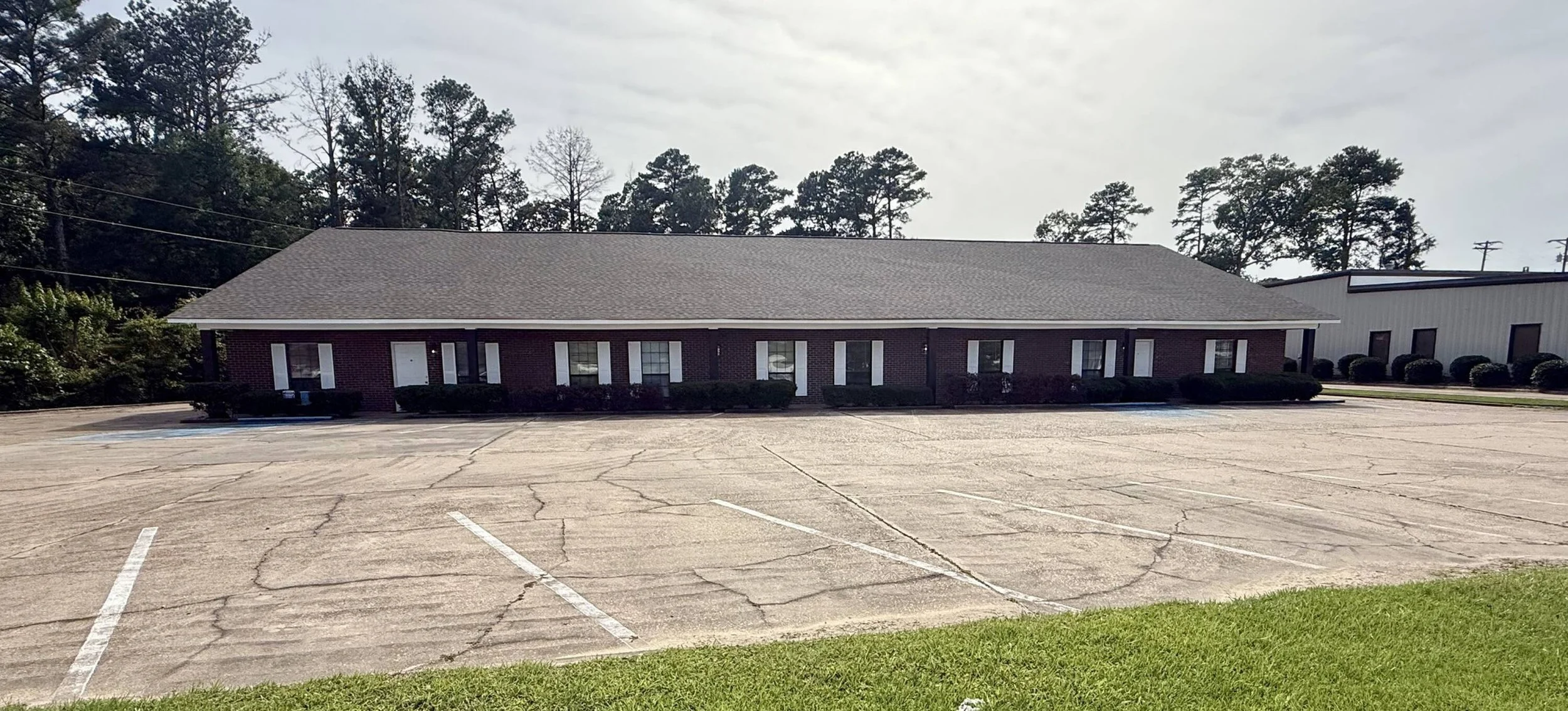 A small, single-story brick building with multiple windows and a gray shingle roof, surrounded by an empty, cracked parking lot and some greenery in the foreground.