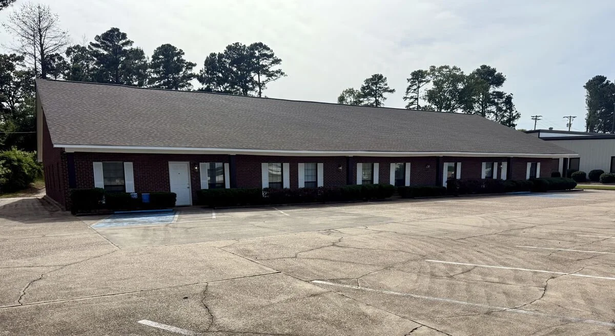 A single-story brick building with multiple units, situated behind an empty parking lot with cracking pavement. There are trees and cloudy skies in the background.