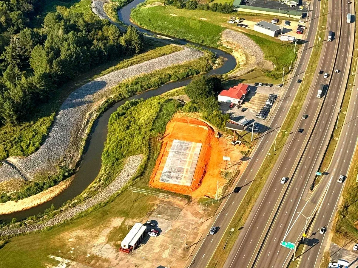 Aerial view of a construction site next to a highway with a river running nearby, and a small building with a red roof.