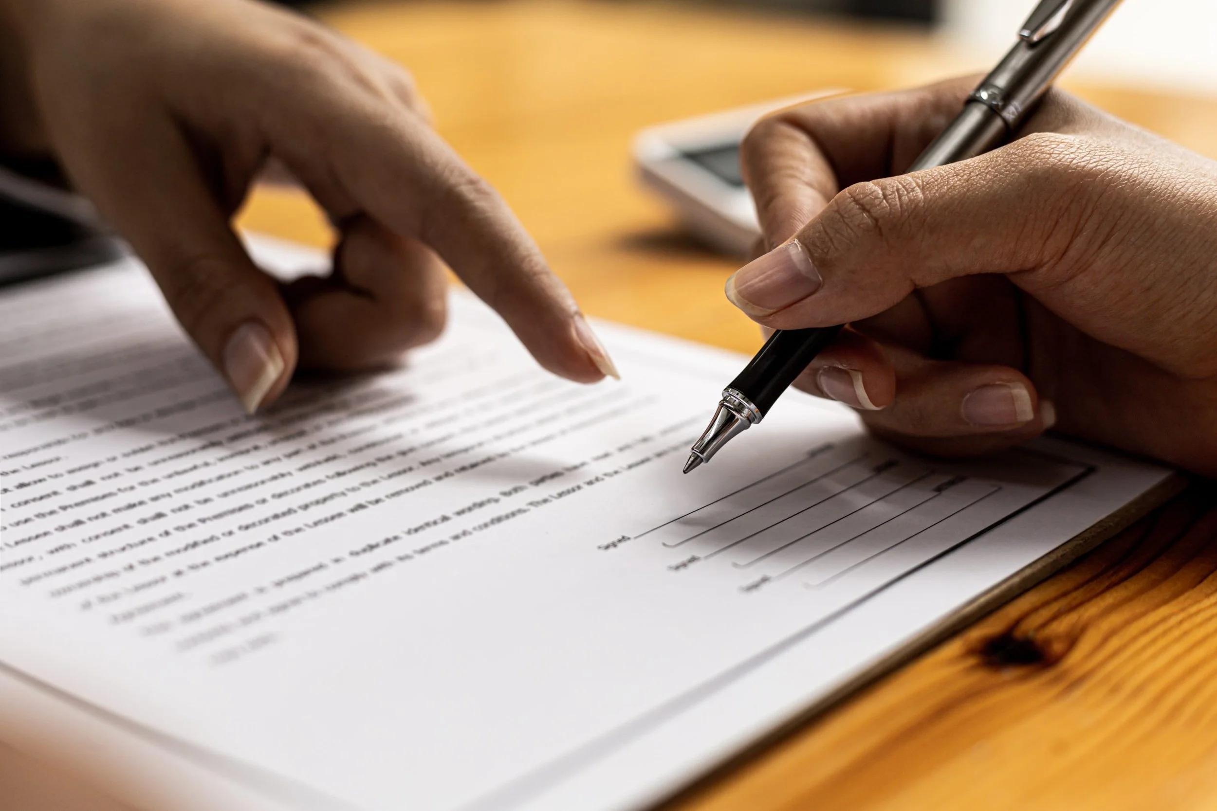 Close-up of a person's hands, one pointing and the other writing with a pen on a document with printed text and a form, placed on a wooden table.