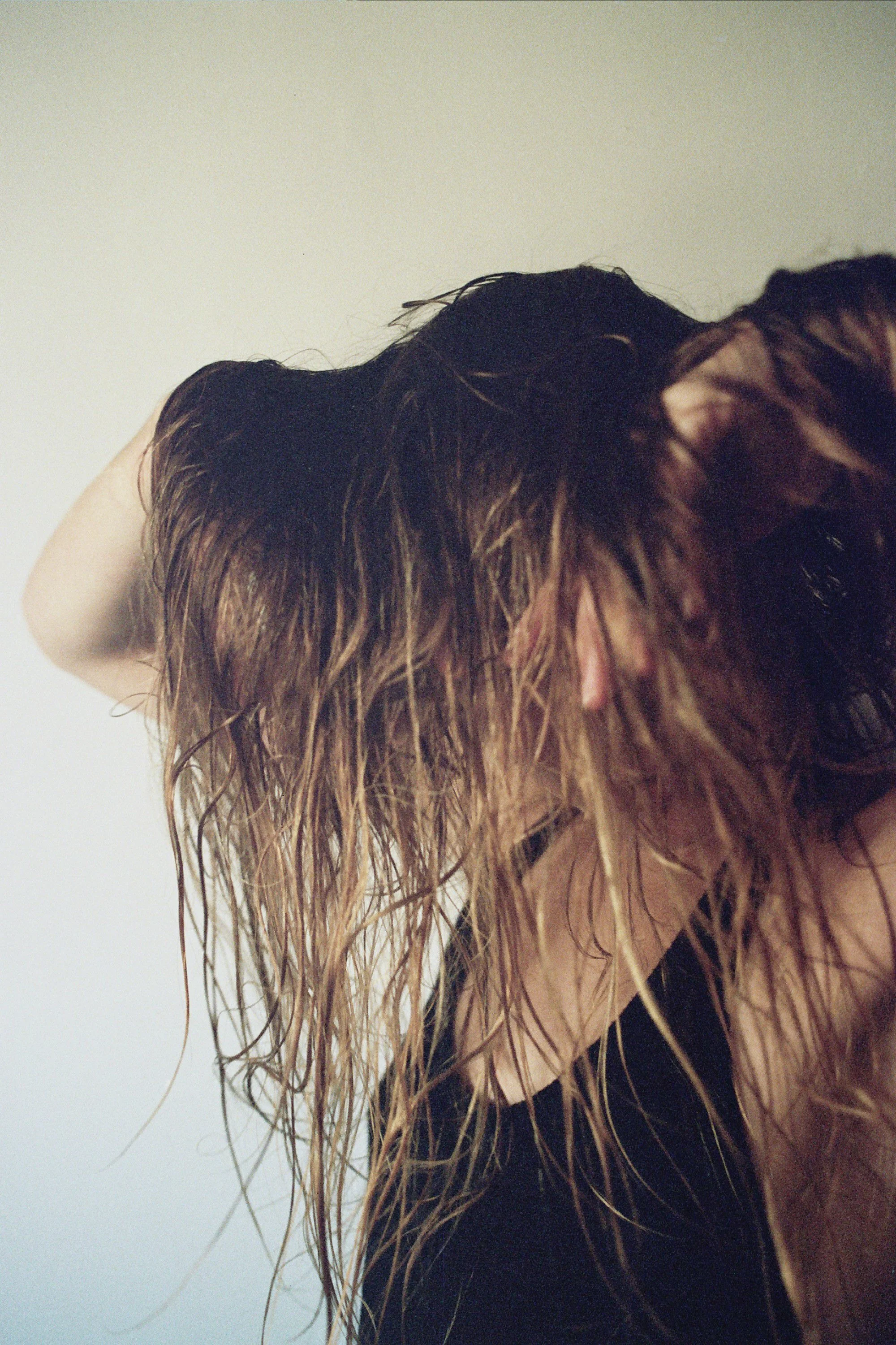 A person with long, wet hair leaning forward, wearing a beige jacket over a black shirt, against a plain white background.