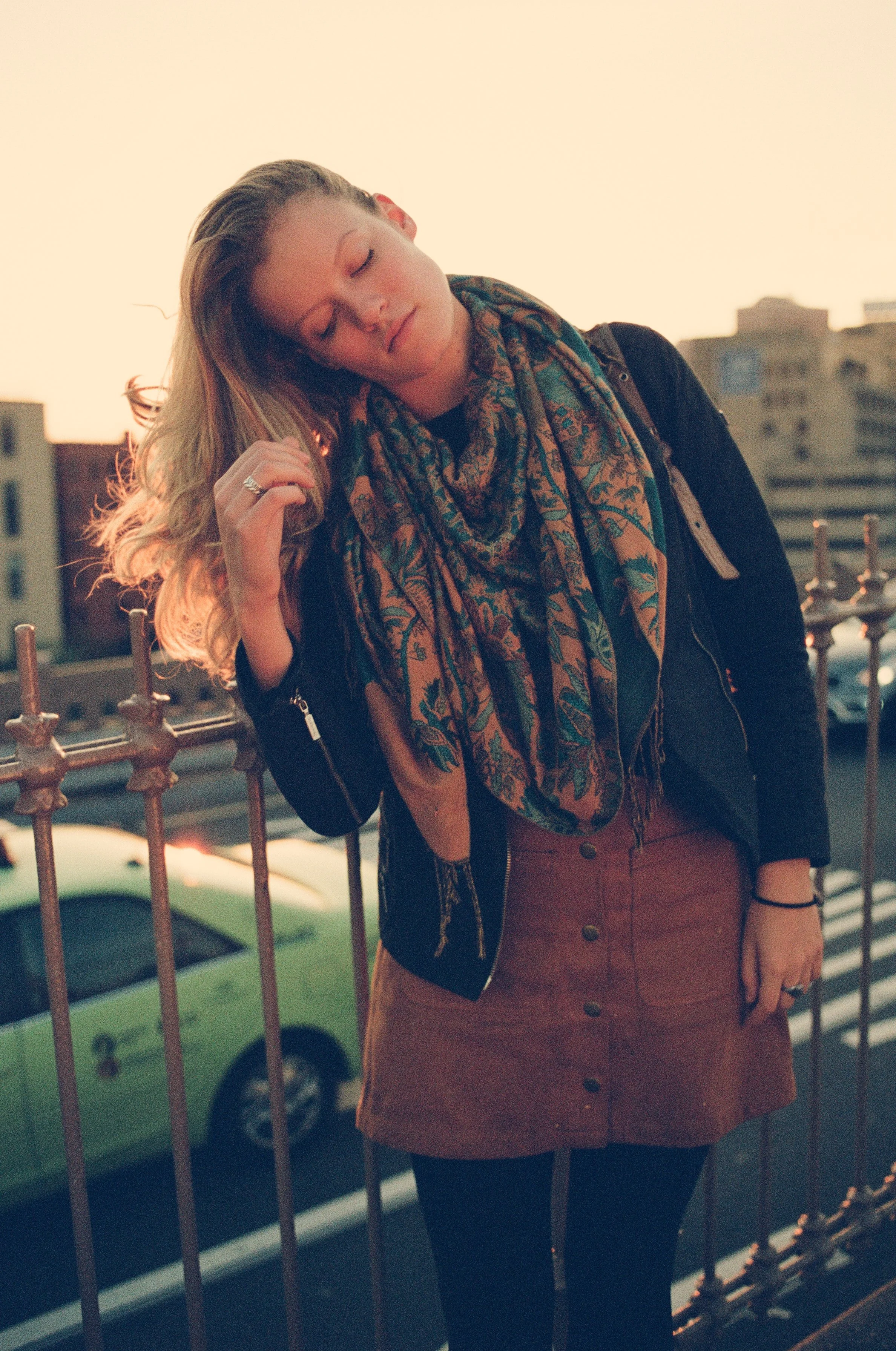 A young woman with long blonde hair standing on a rooftop or balcony, leaning against a metal railing, with her eyes closed and a peaceful expression, during sunset or golden hour. She is wearing a black jacket, a colorful scarf, a brown skirt, and b
