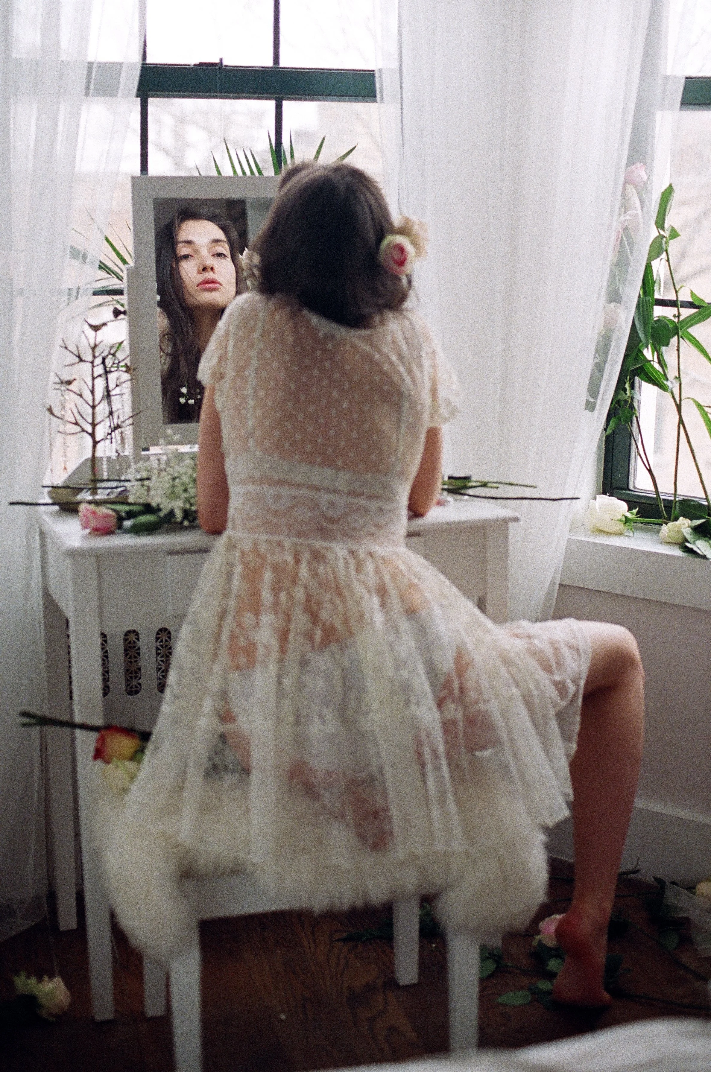 Young woman in vintage dress sitting at vanity, looking at her reflection in a mirror, surrounded by flowers and plants in a softly lit room.