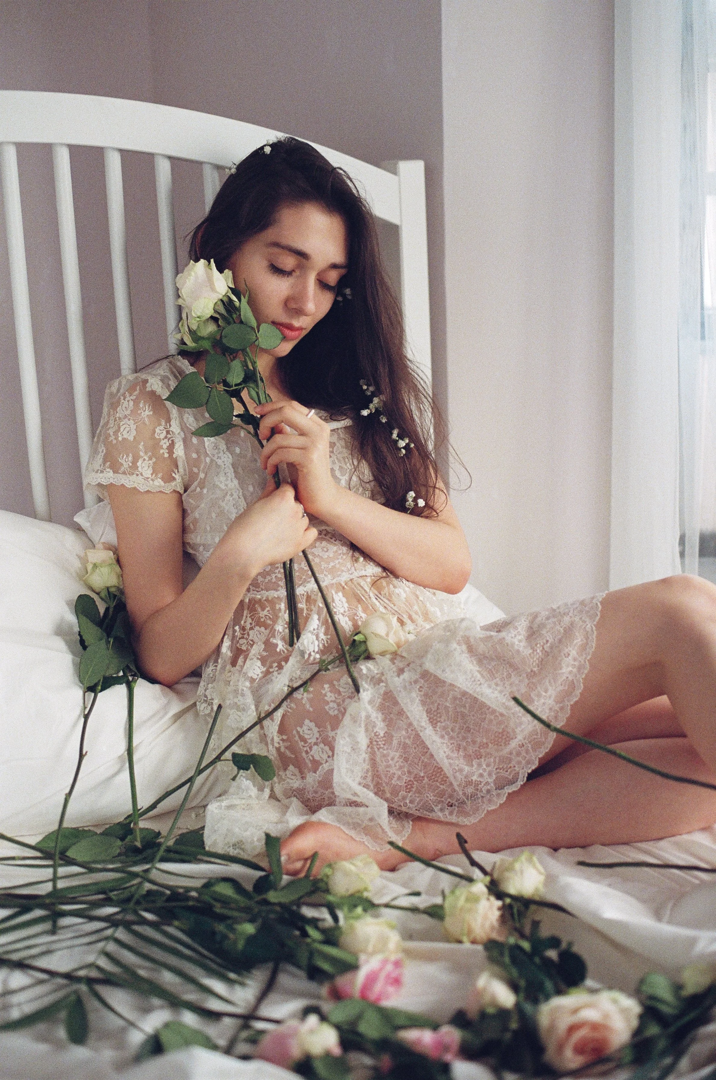 A young woman in a lace dress sitting on a bed surrounded by white roses, holding one rose near her face with a serene expression.