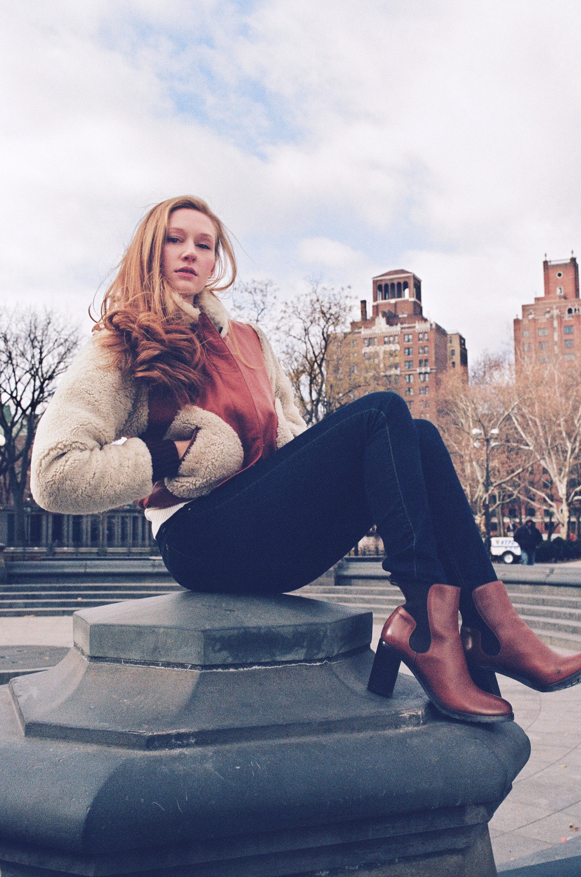 A woman with long red hair sitting on a stone ledge outdoors, wearing a beige and red jacket, black pants, and brown ankle boots. In the background, leafless trees and tall buildings are visible under a partly cloudy sky.