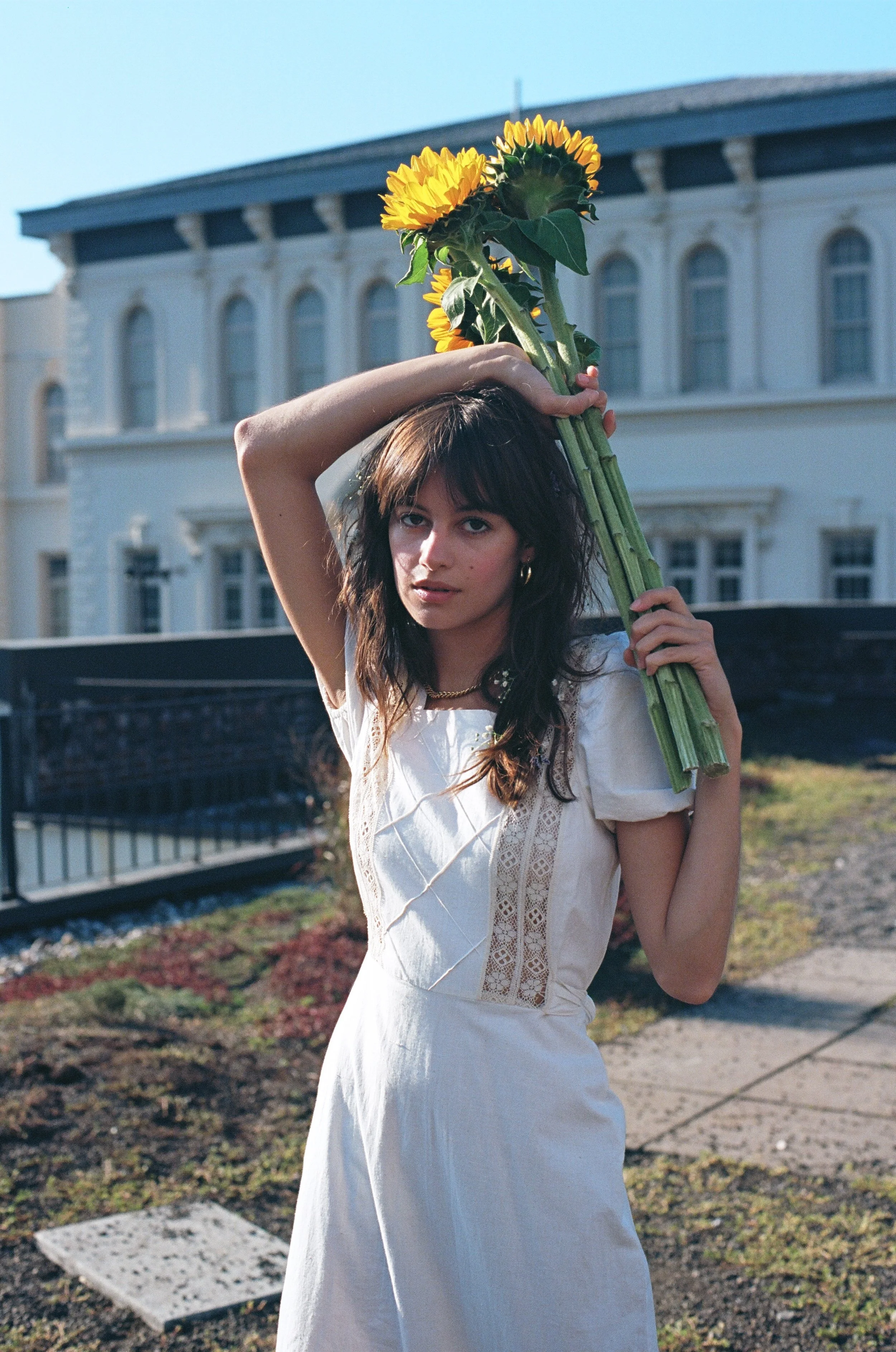A woman holding a bouquet of sunflowers over her shoulder outdoors in front of a light-colored building.