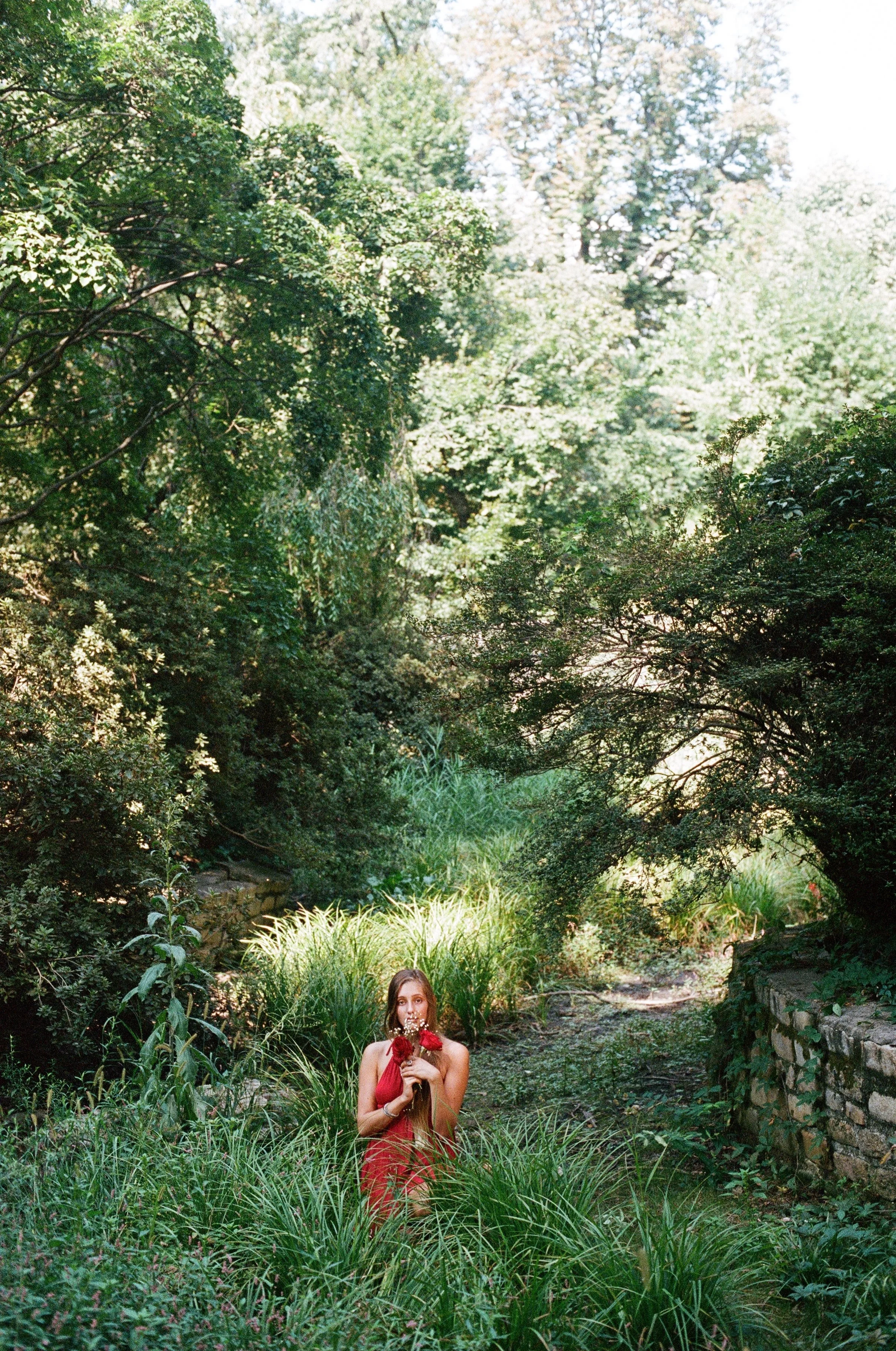 A woman in a red dress holds a bouquet of red flowers while standing among tall grass and lush greenery in a garden.