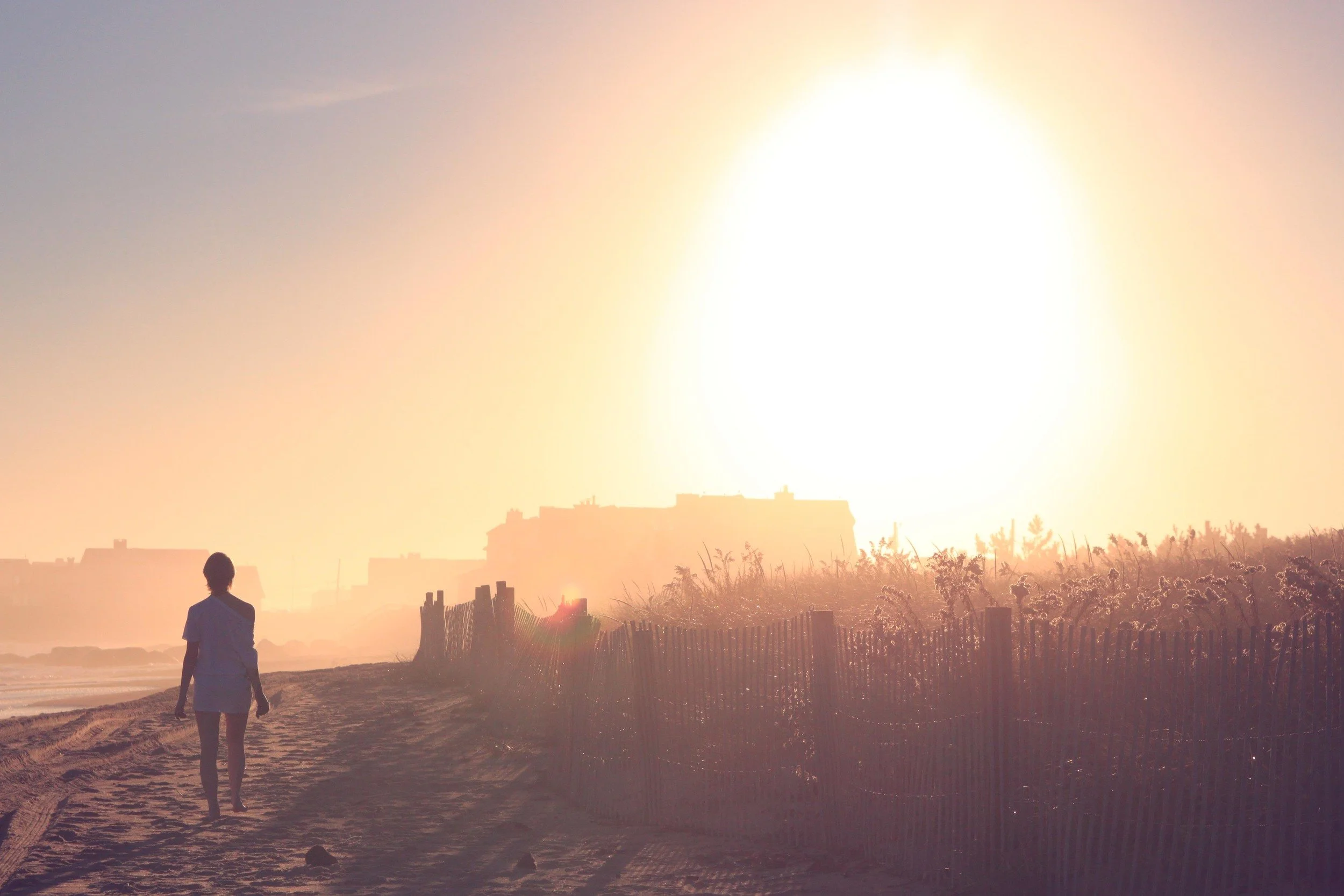 Person walking on a sandy beach with a fence, with the sun setting or rising in the background.