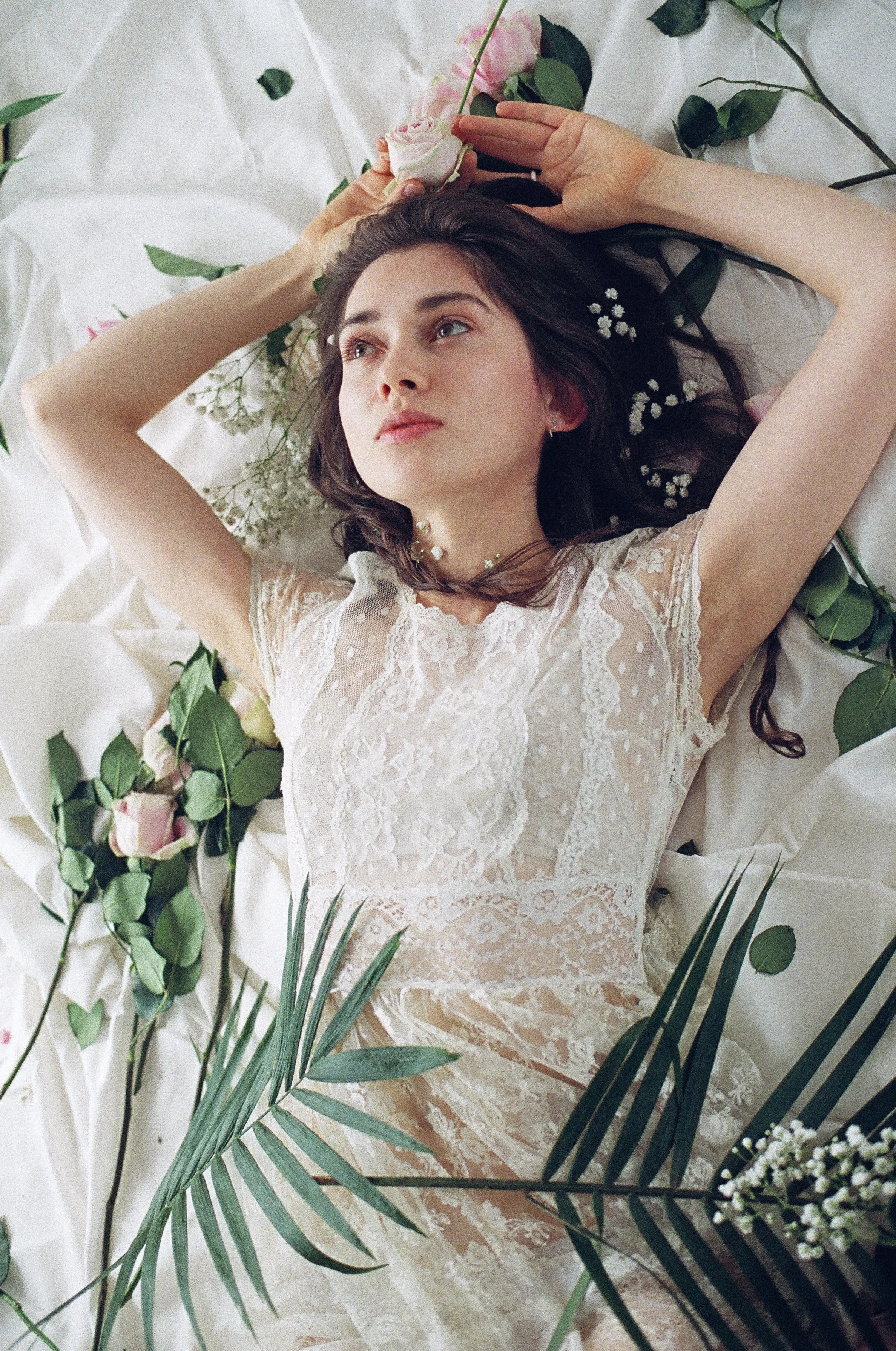 A woman with dark hair lying on white fabric surrounded by flowers and green leaves, wearing a lace dress.