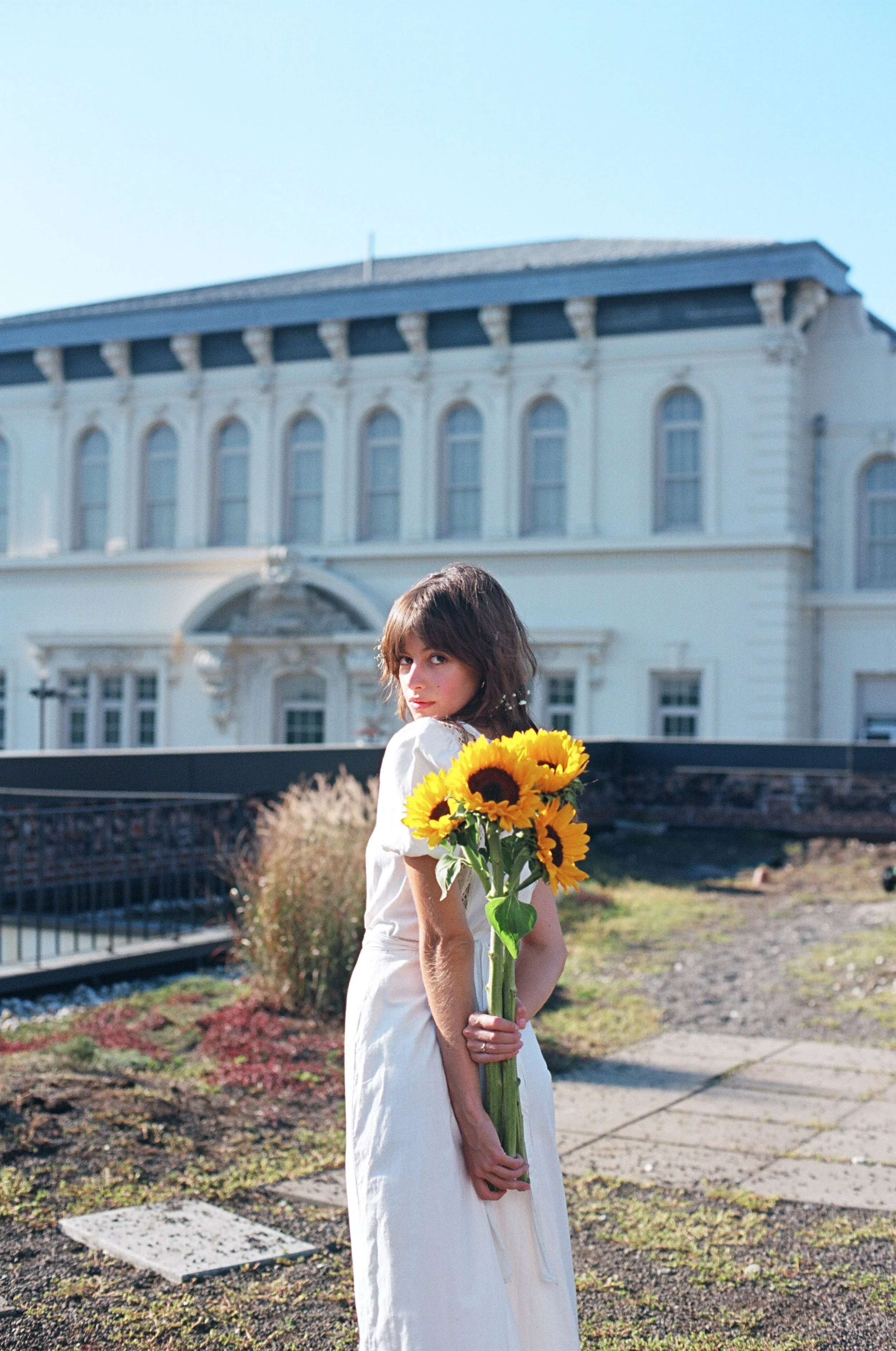 A woman in a white dress holding a bouquet of sunflowers outdoors with a large historic building in the background.