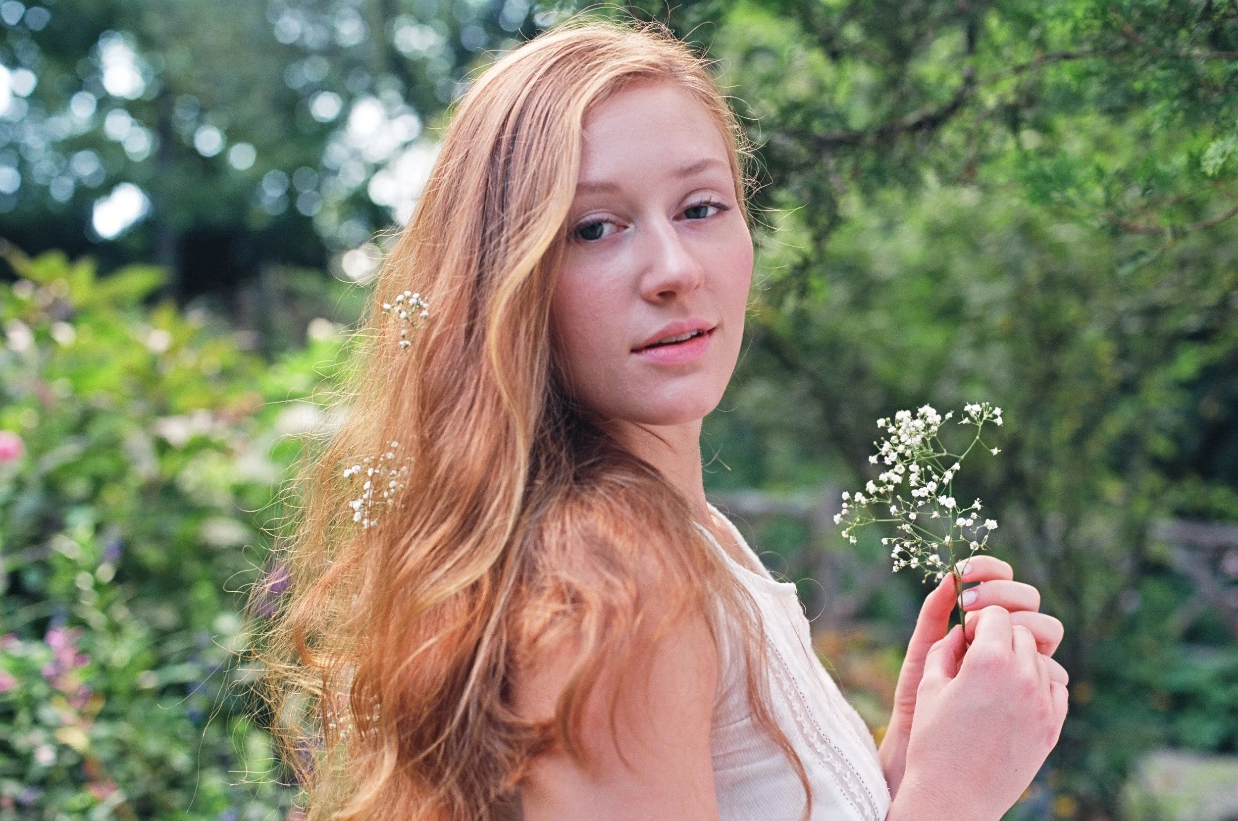 A young woman with long, wavy red hair holding a small white flower, standing outdoors in a lush green garden.