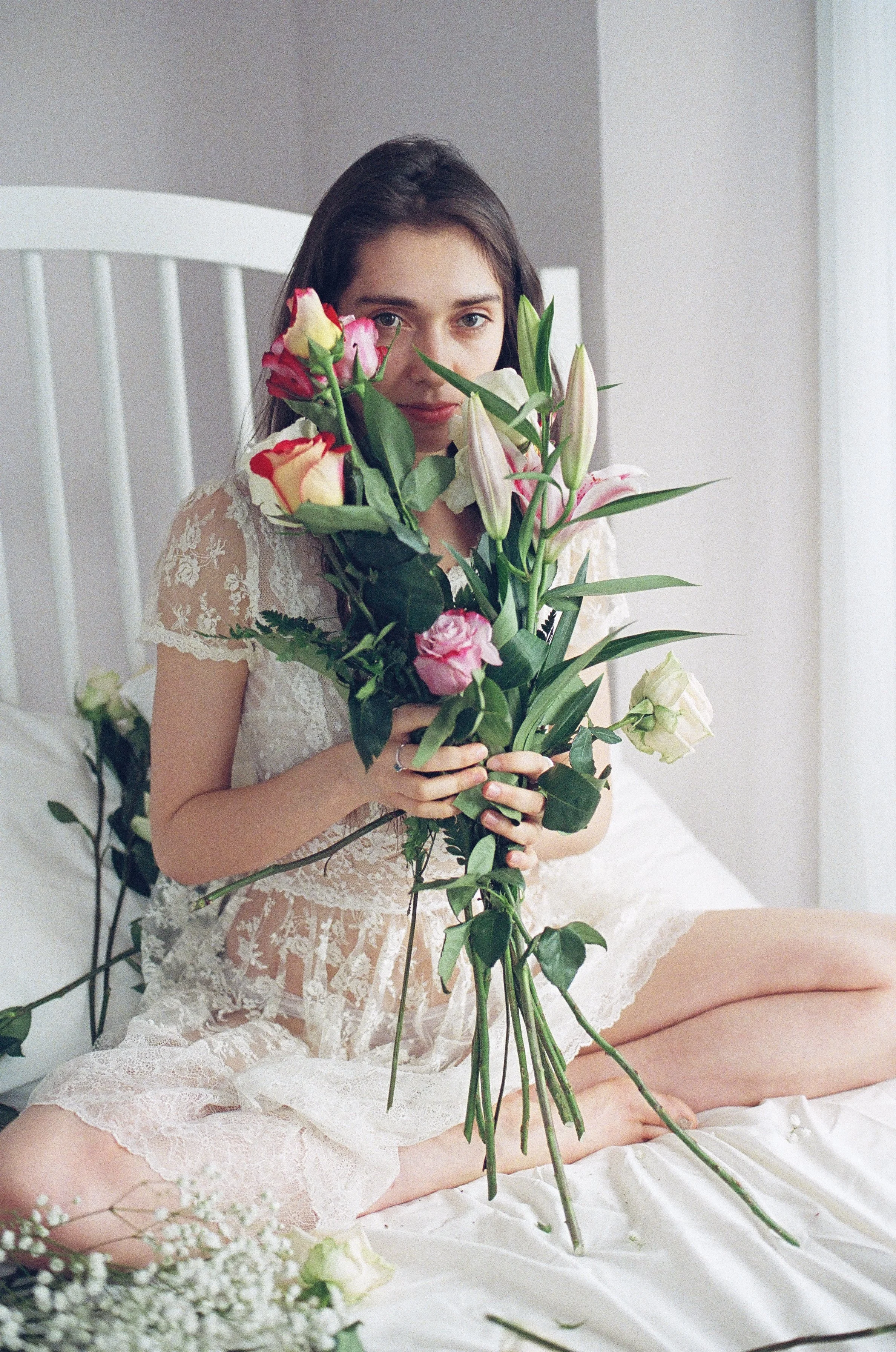 A young woman in lace dress sitting on bed, holding a large bouquet of pink, white, and yellow flowers, with some flowers and petals scattered around her on the bed.