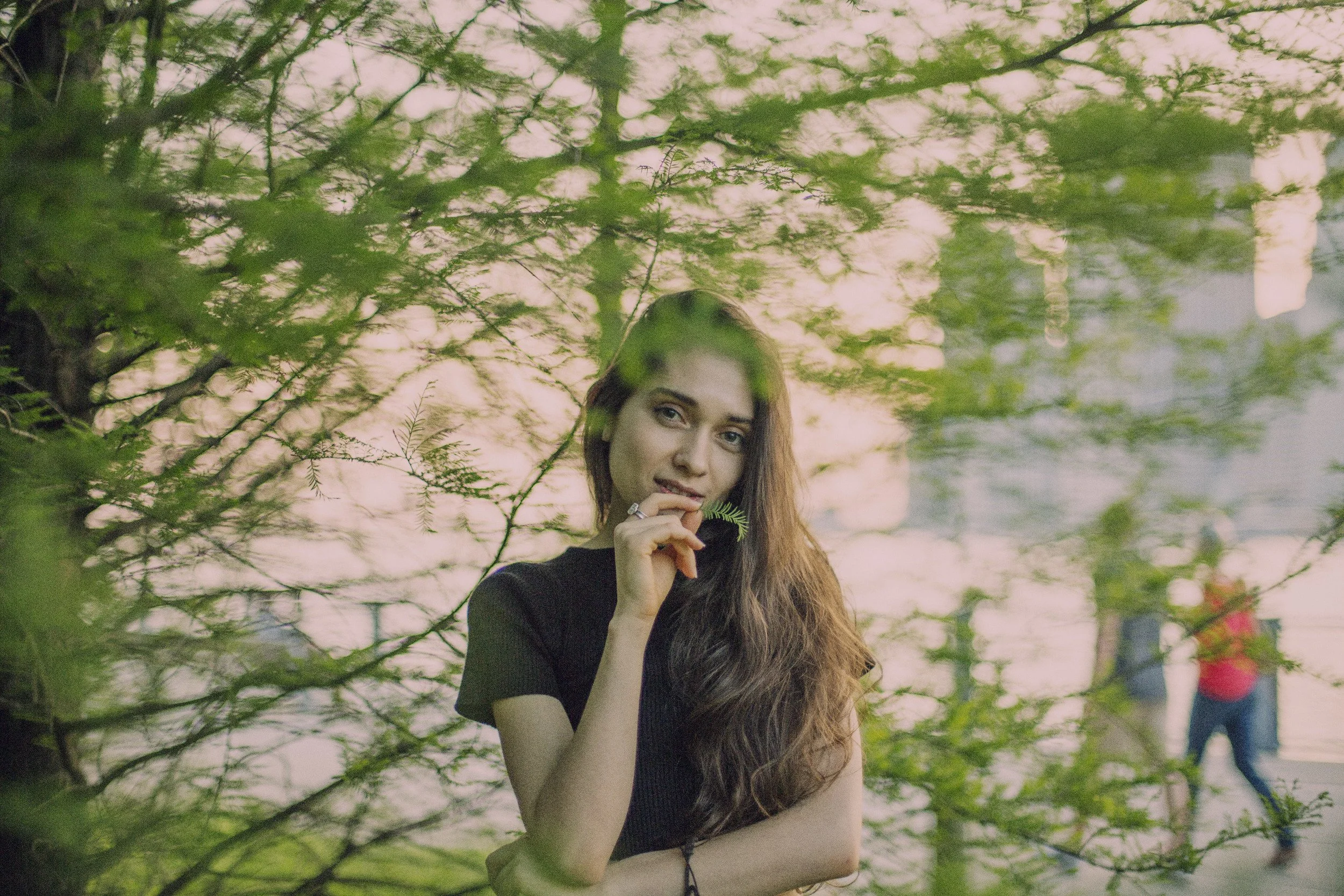 A young woman with long brown hair holding a small green leaf near her face, standing outdoors among trees with a body of water and two people walking in the background at sunset.