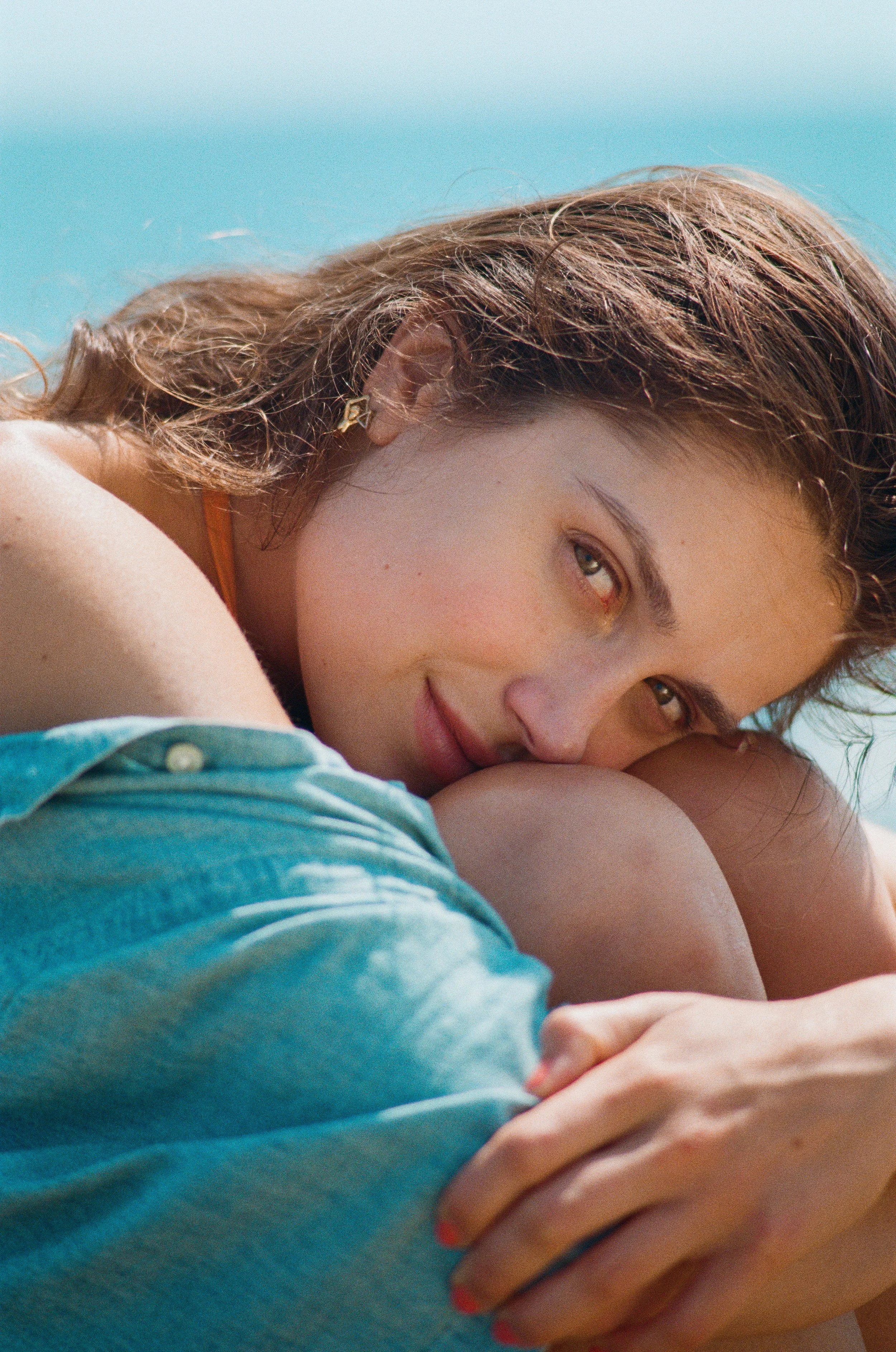 A woman with wavy brown hair resting on her knees, looking at the camera with a gentle smile, outdoors with a blue sky background. Photo edited by Mary Perrino.