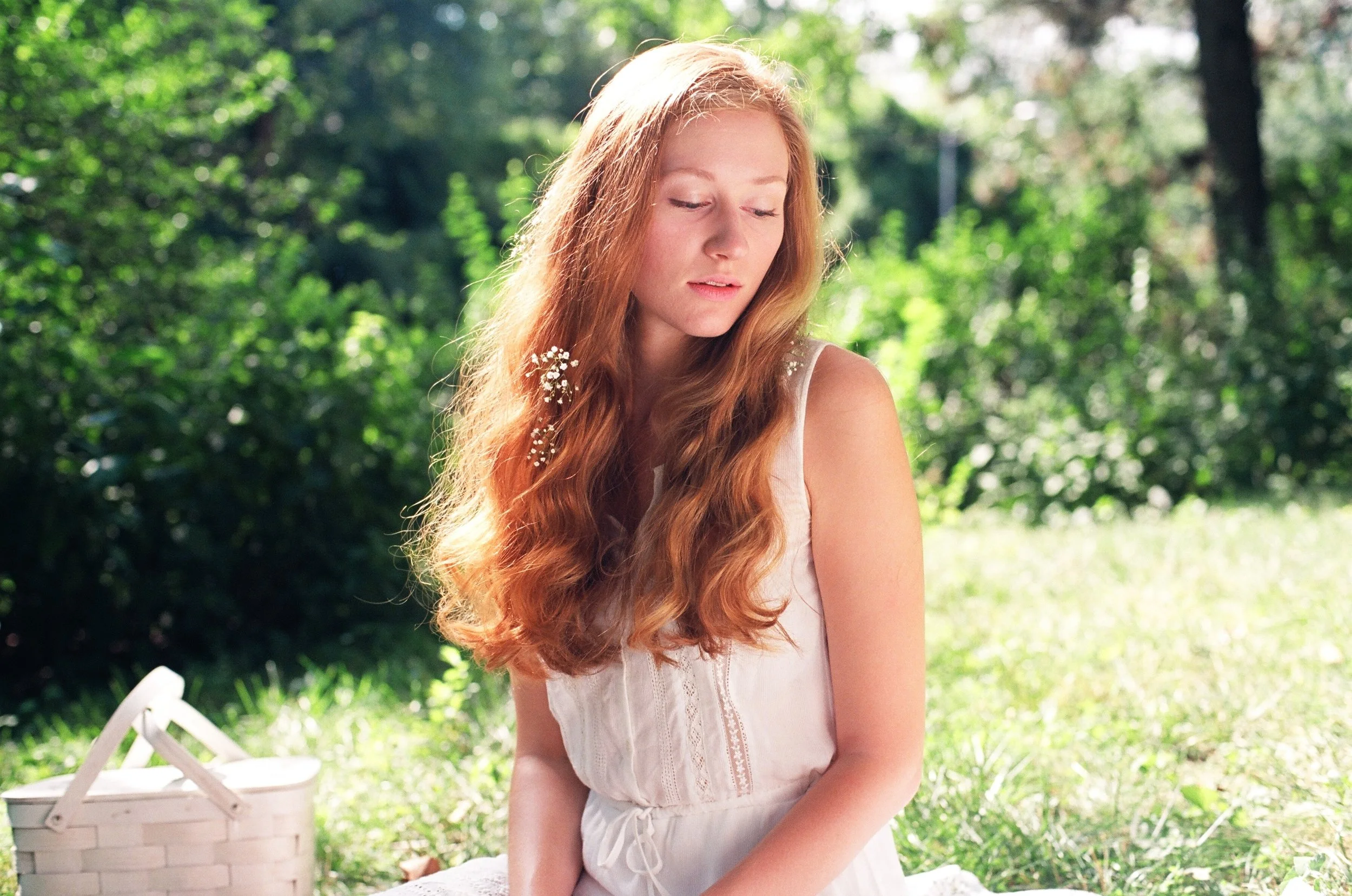 A young woman with long red hair sits outdoors in a lush green setting with sunlight filtering through the trees. She is wearing a white sleeveless dress and appears to be lost in thought. There is a white picnic basket next to her.