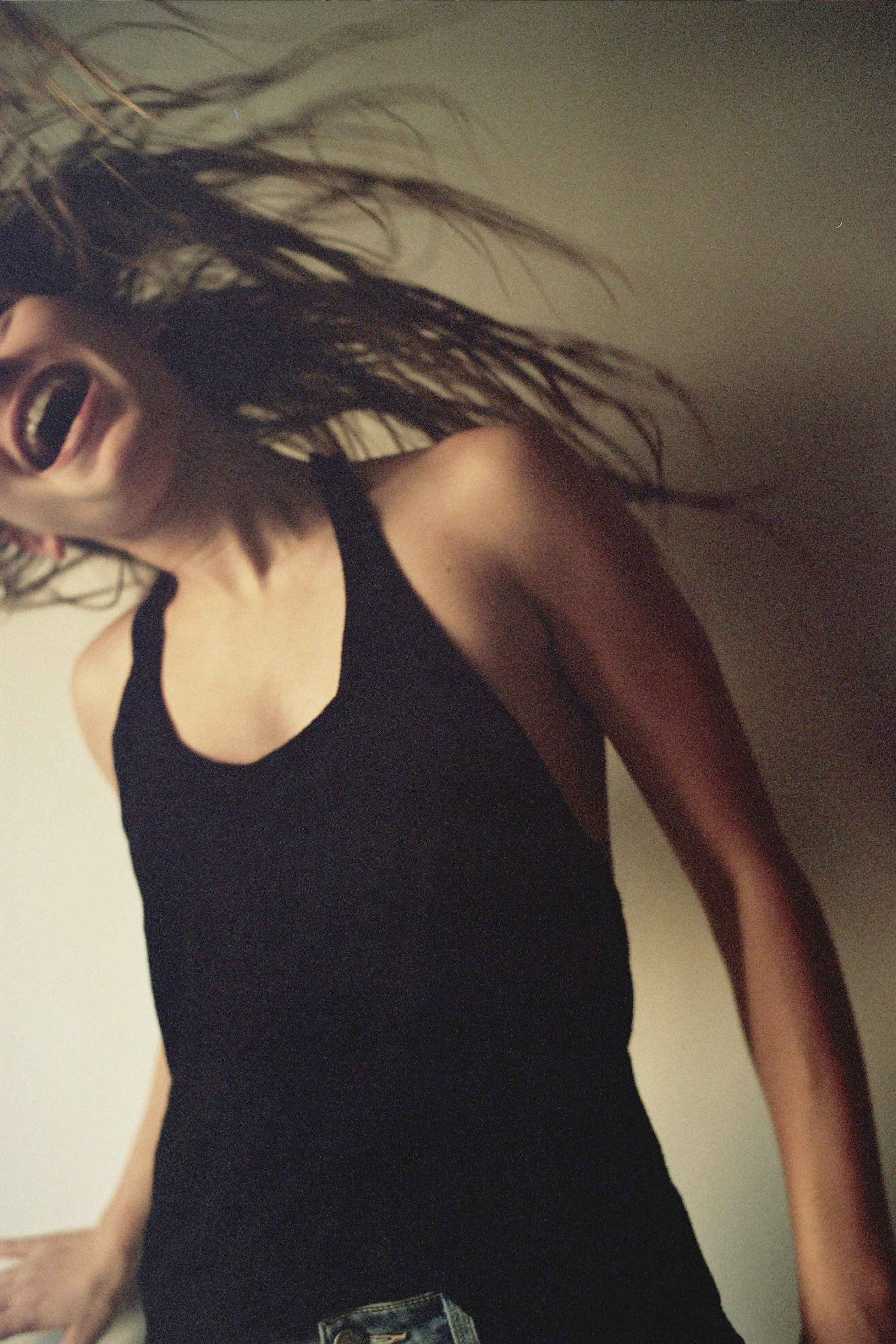 A young woman with long, wavy hair wearing a black tank top, laughing and leaning against a wall.