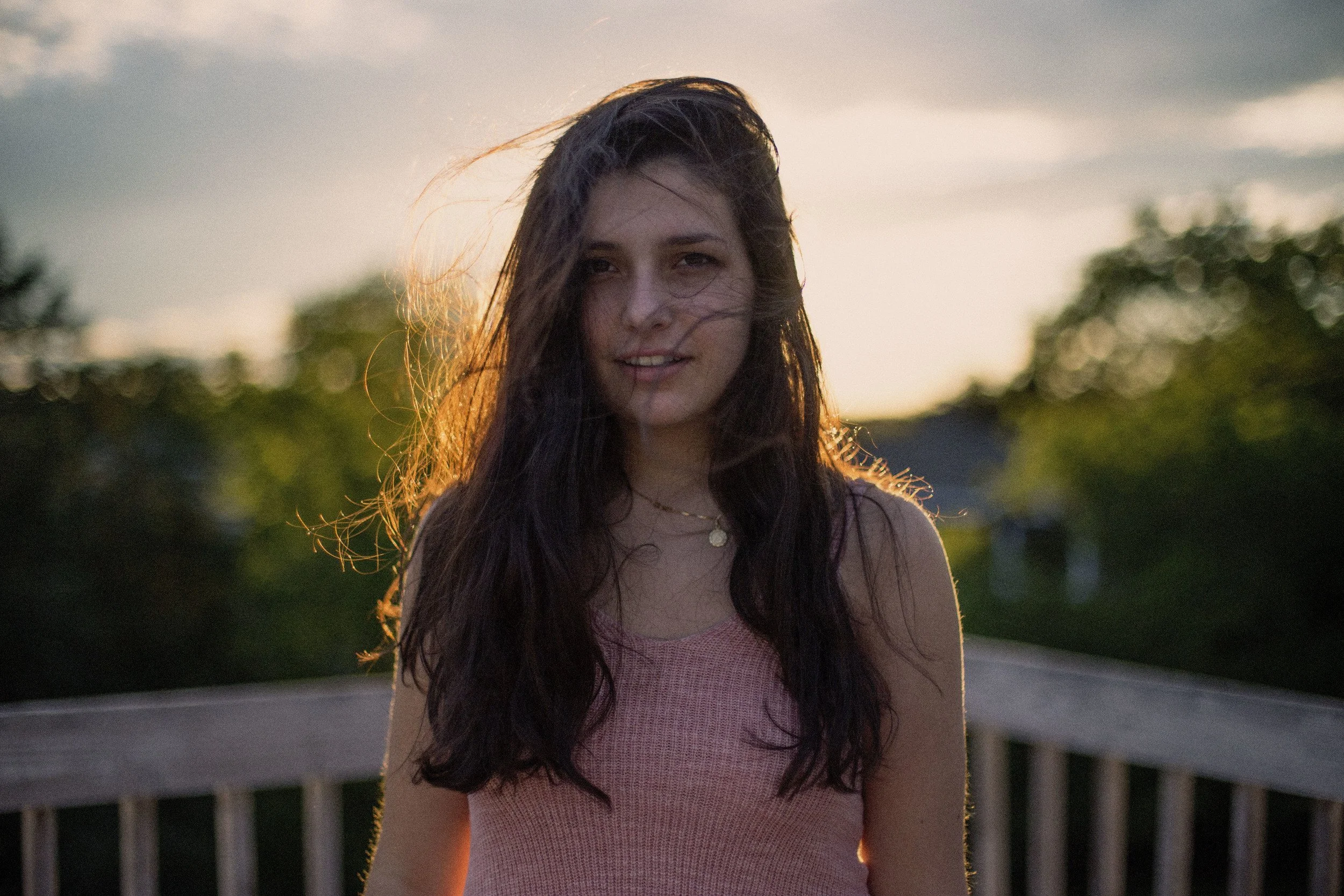 A young woman with long dark hair standing outdoors during sunset, with trees and a fence in the background.