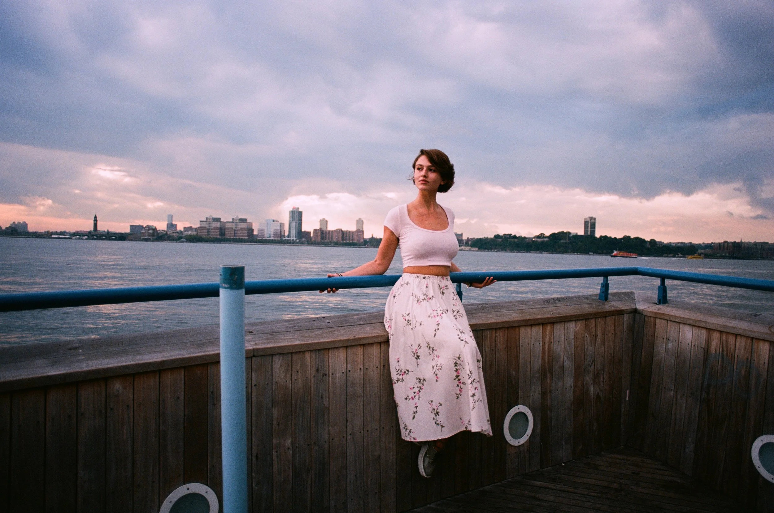 A woman in a white crop top and long floral skirt leaning on the railing of a wooden dock overlooking a body of water with a city skyline and cloudy sky in the background.