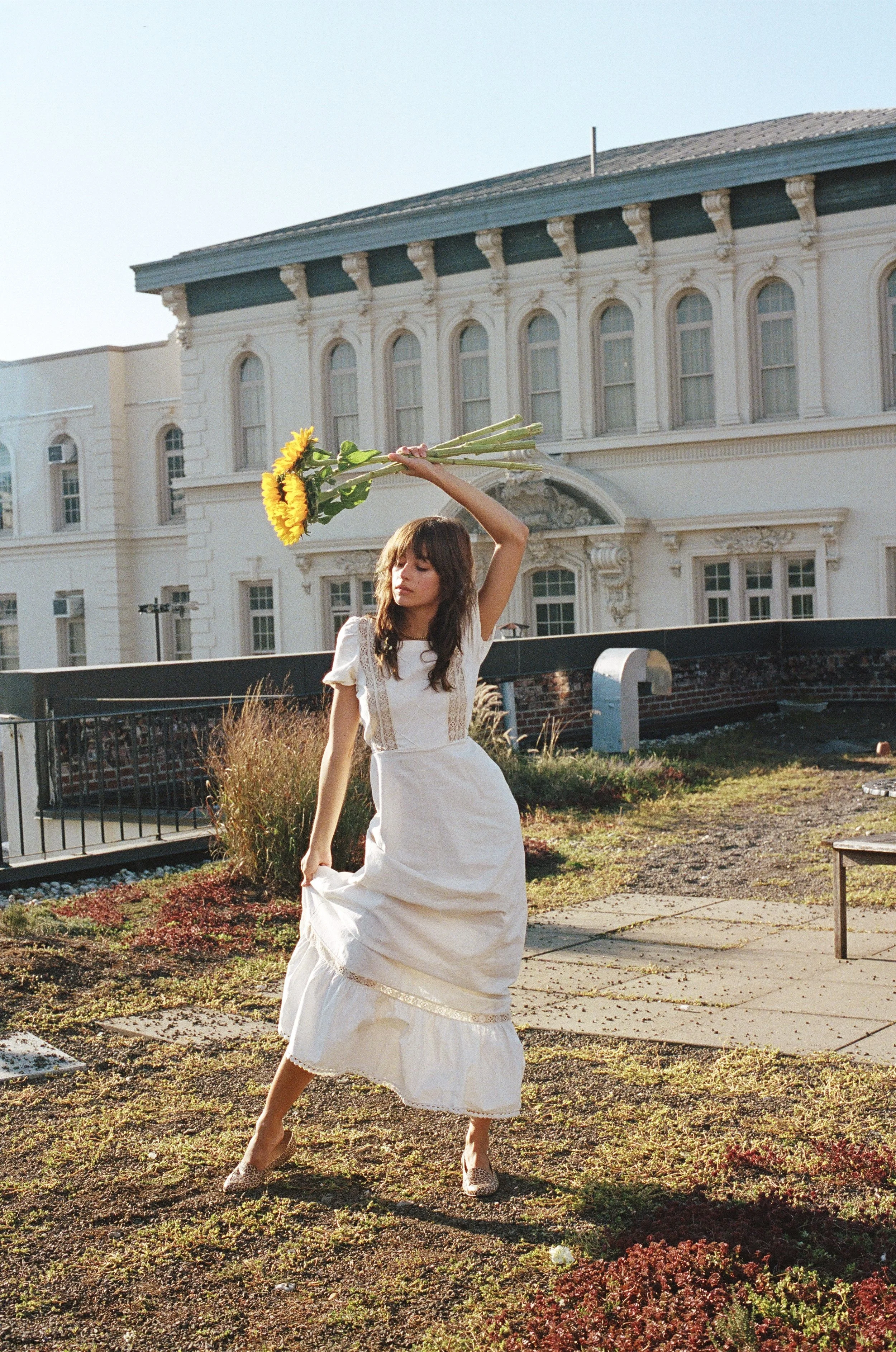 A woman in a white dress holding a bouquet of sunflowers above her head on a rooftop with a large ornate white building in the background.