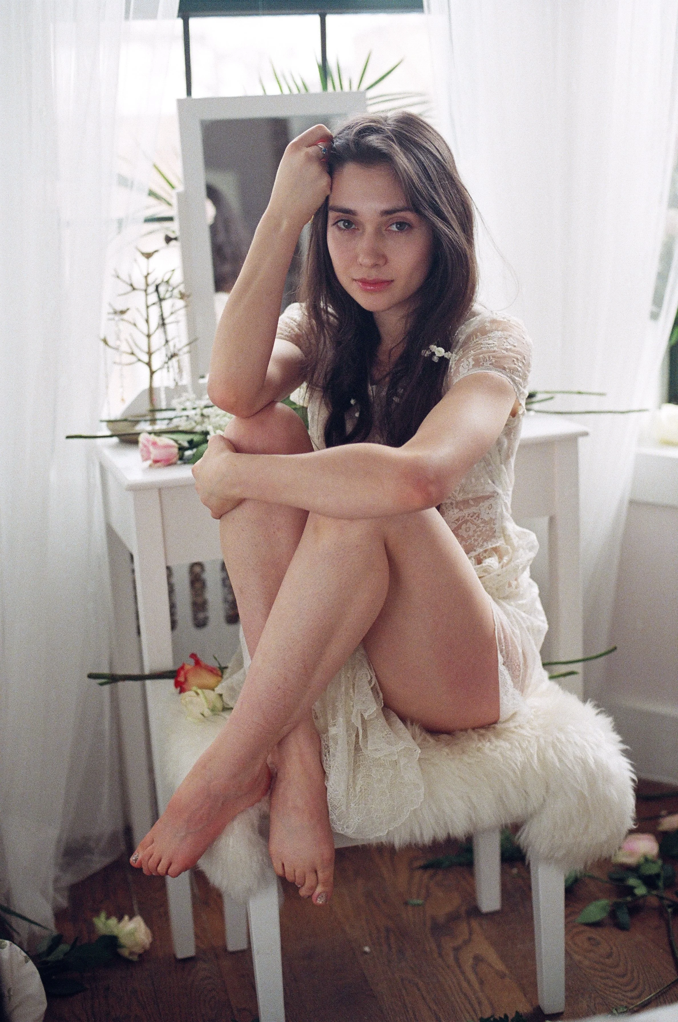 Young woman with long dark hair sitting on a white furry stool in front of a dresser with a mirror, surrounded by flowers and plants, in a brightly lit room.