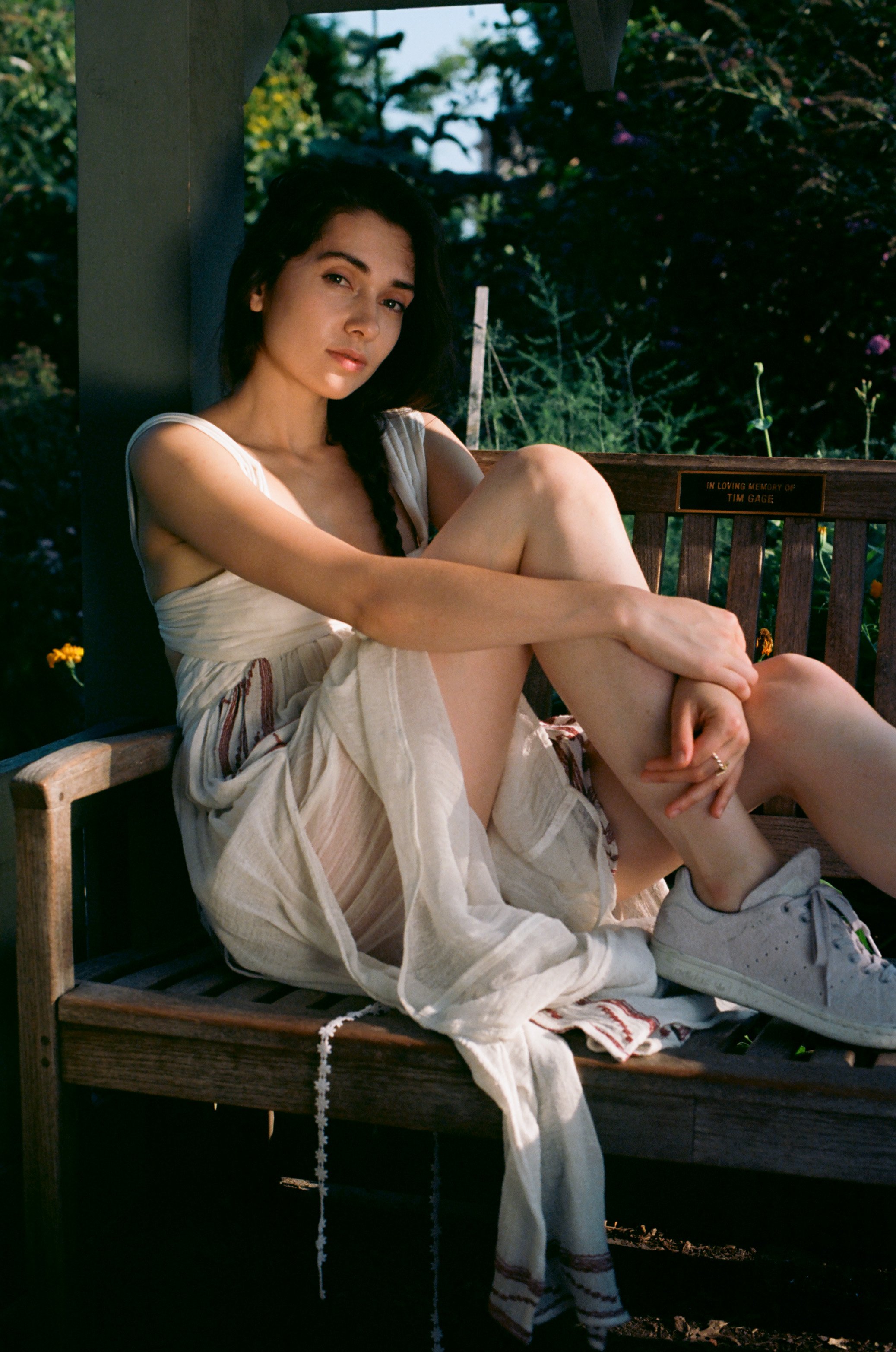 Woman sitting on wooden bench with greenery in the background, wearing a light-colored dress and sneakers.