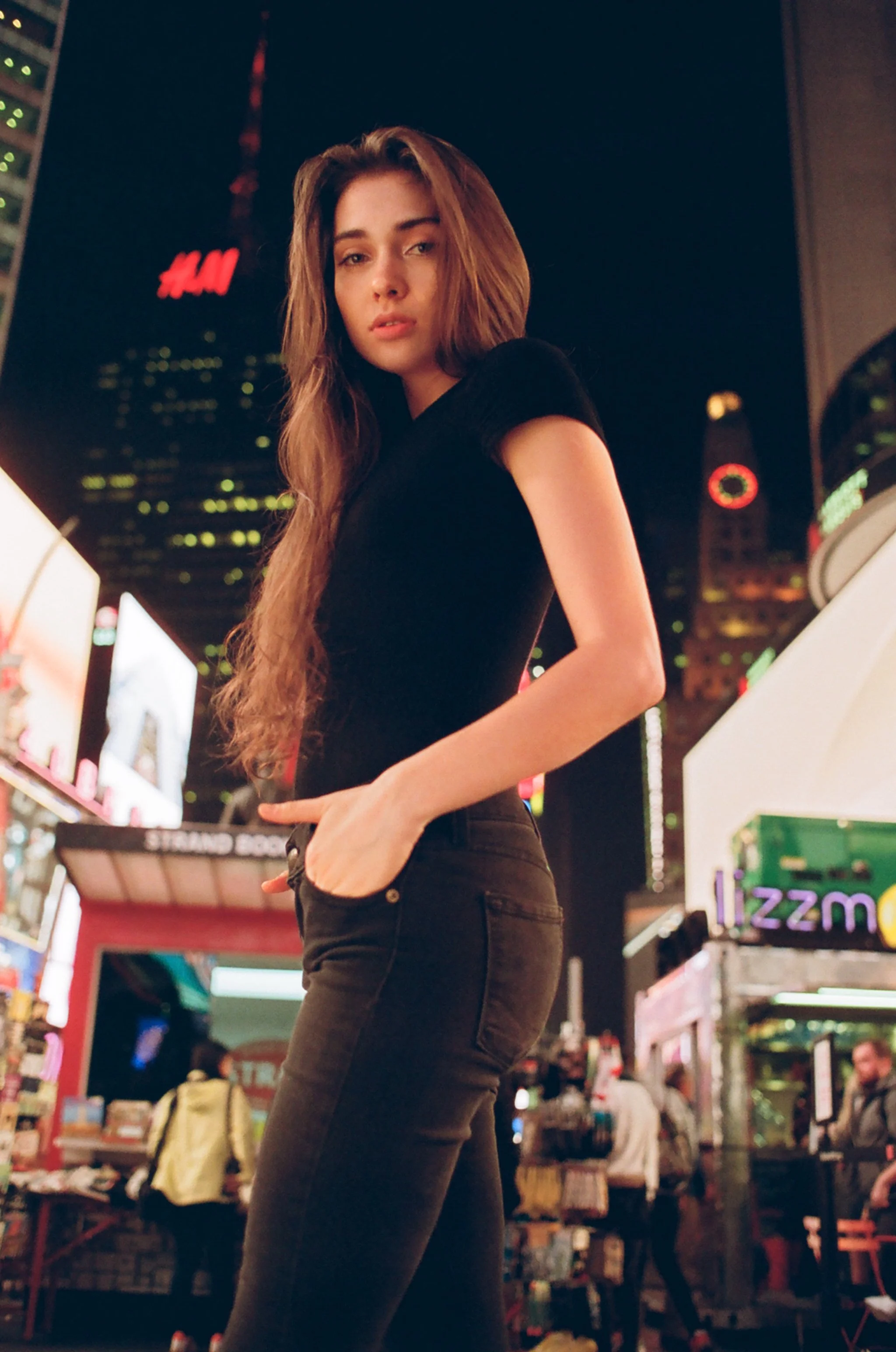 Young woman standing at night in Times Square with bright lights and billboards in the background, wearing a black top and black jeans, with her hand in her pocket.