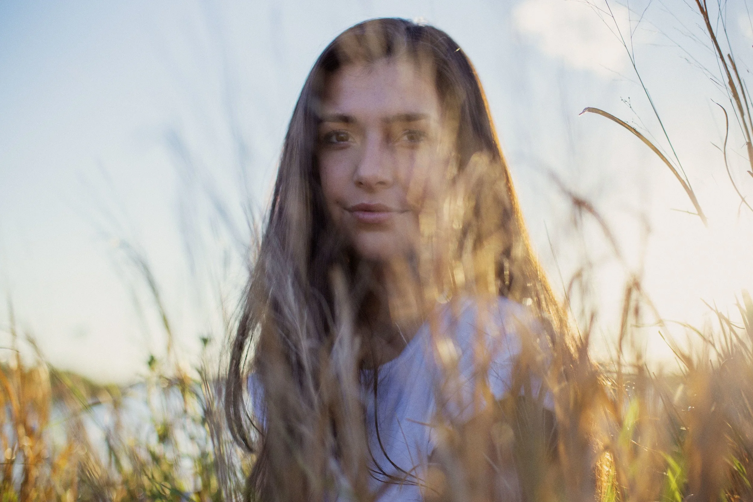 A young woman with long brown hair standing in a field of tall grass during sunset.