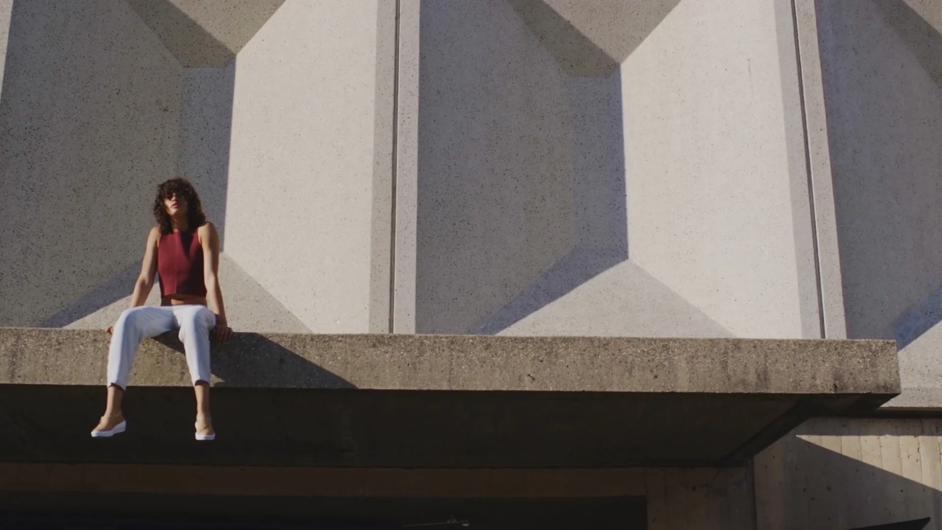 A woman with curly hair sitting on the edge of a concrete structure in front of a building with large geometric shadows and patterns.