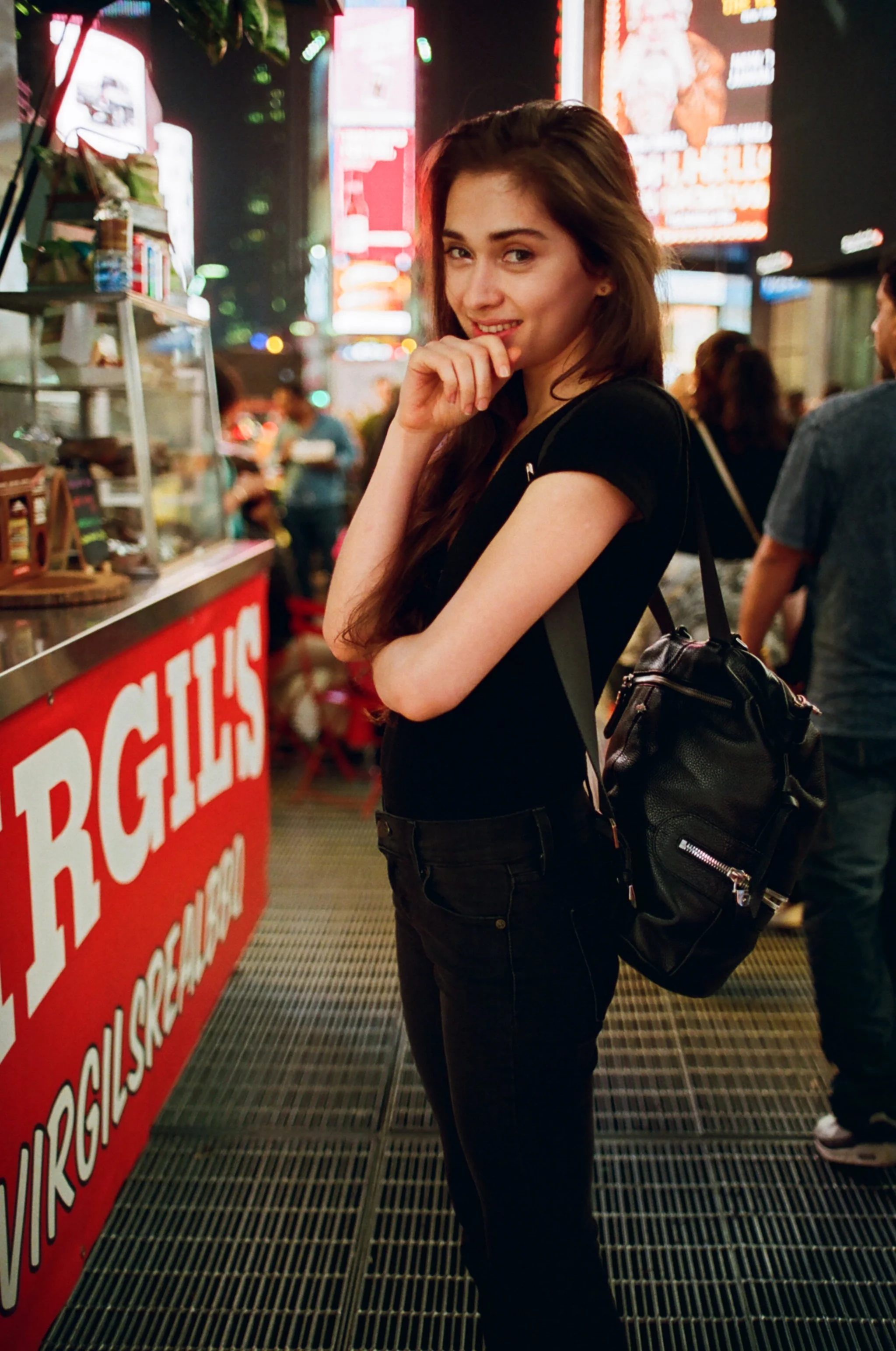 A young woman with long brown hair and a black t-shirt standing in front of a street food stand at night, smiling and looking at the camera, with neon lights and other people in the background. Photo edited by Mary Perrino.