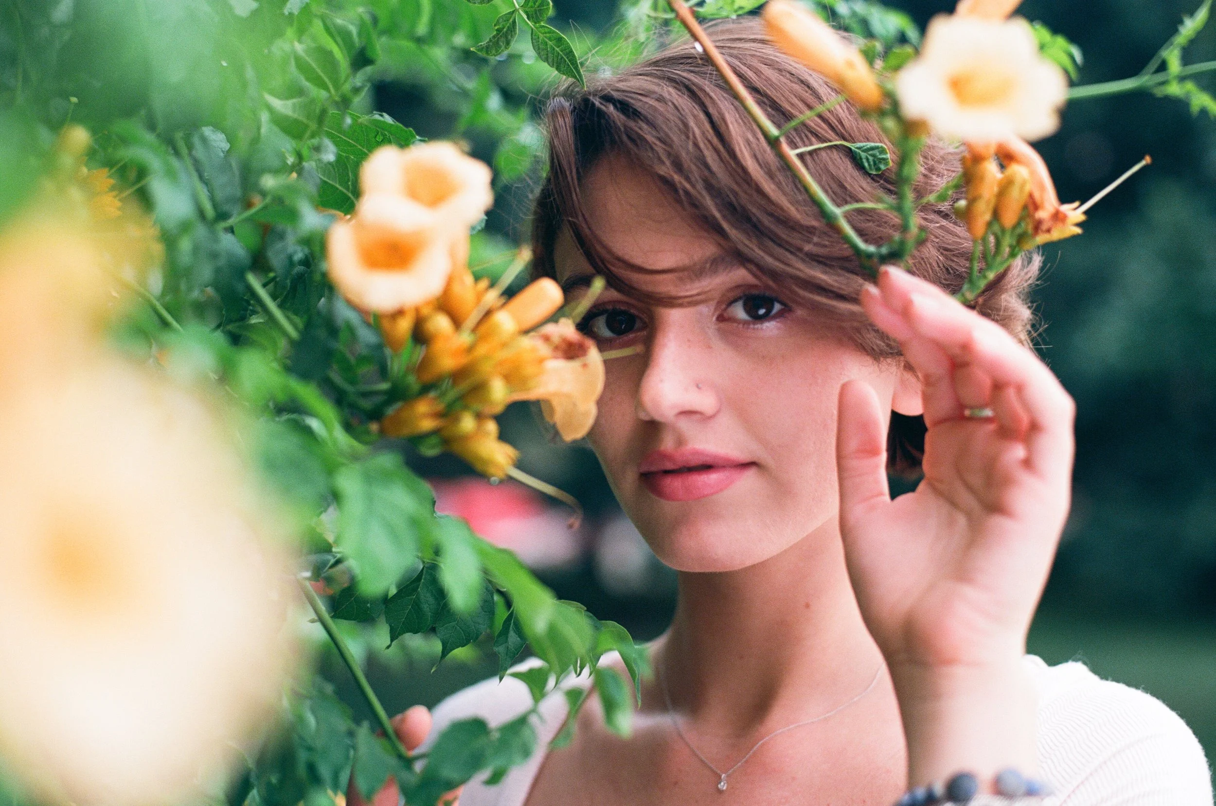 A woman with short brown hair and light skin looks through bright orange and yellow flowers and green leaves, holding a stem close to her face.