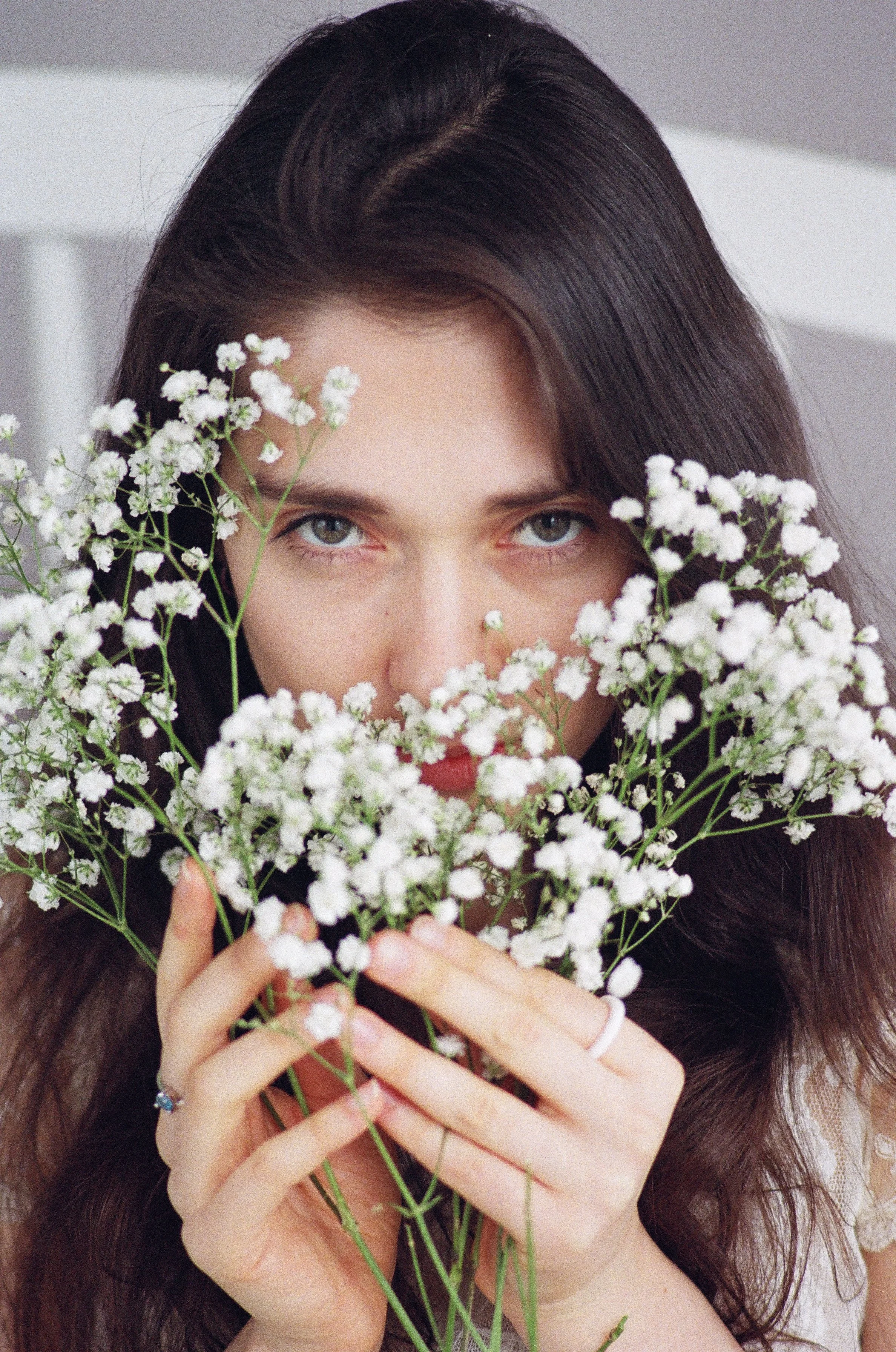 A woman with long dark hair holding a bouquet of white flowers, partially covering her face, looking directly at camera.