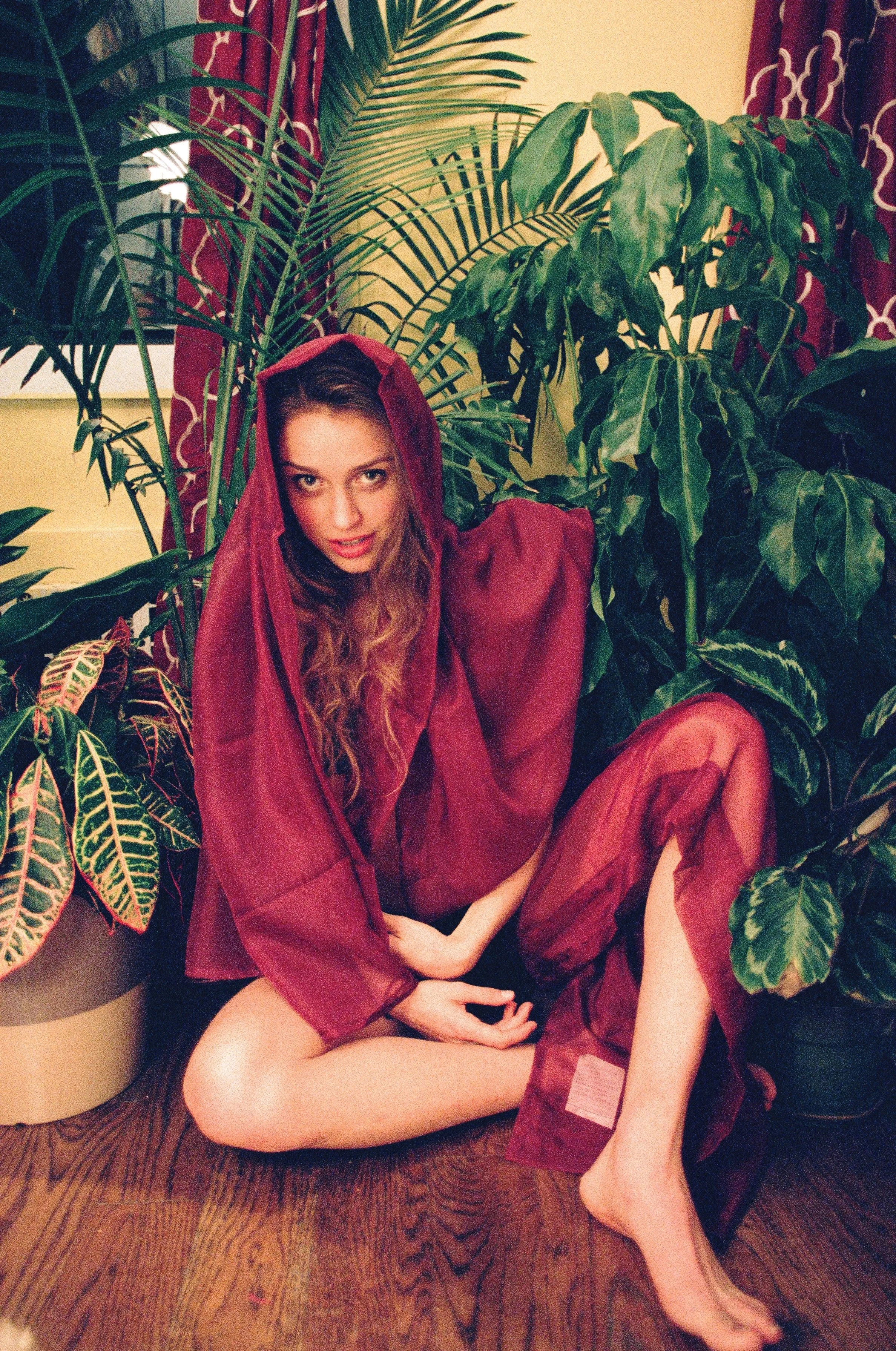 A woman sitting on the floor surrounded by large green houseplants, wearing a red hooded robe with sheer fabric, with a red curtain in the background.