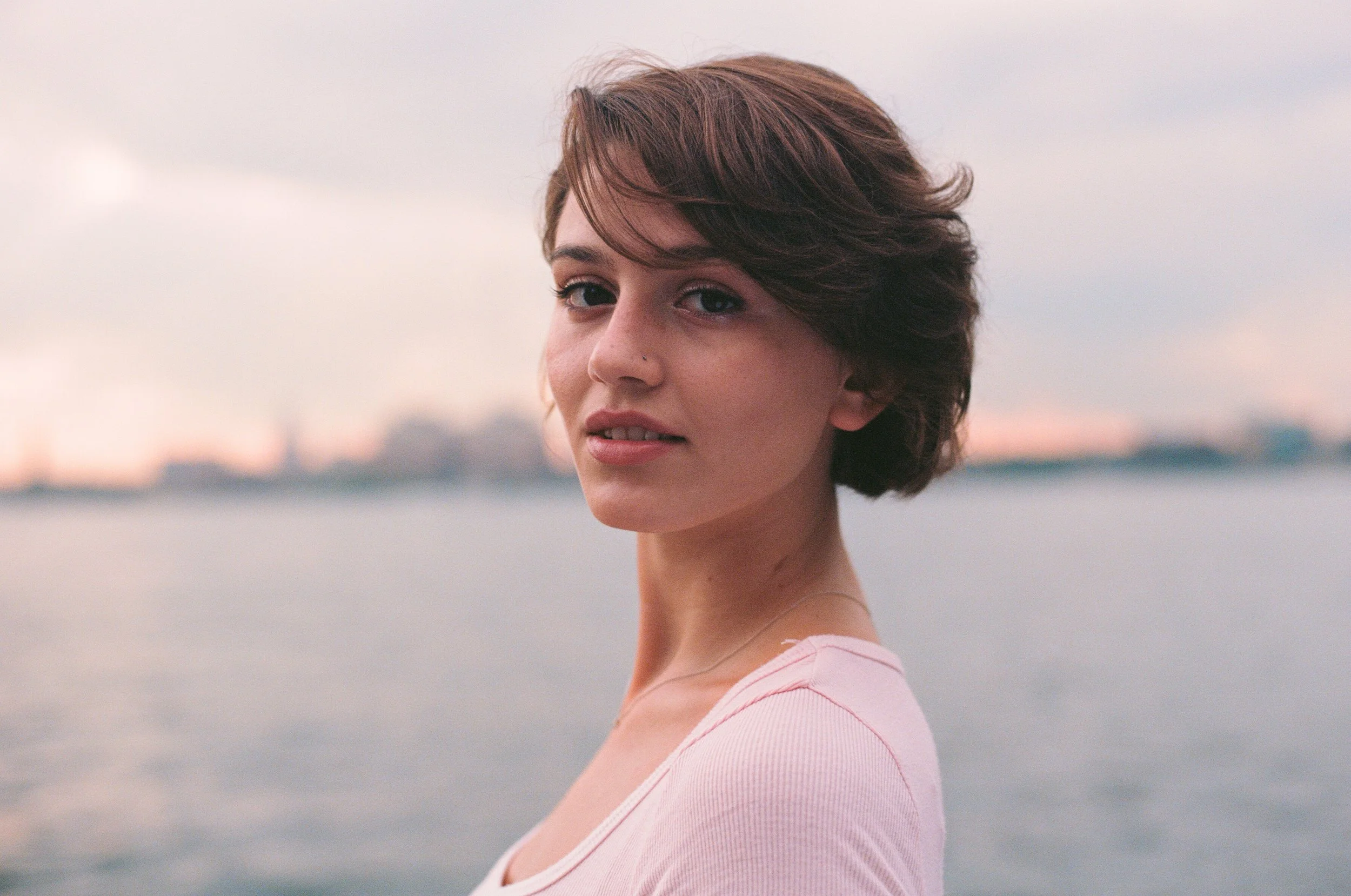 A young woman with short, wavy brown hair standing near a body of water during sunset or dusk.