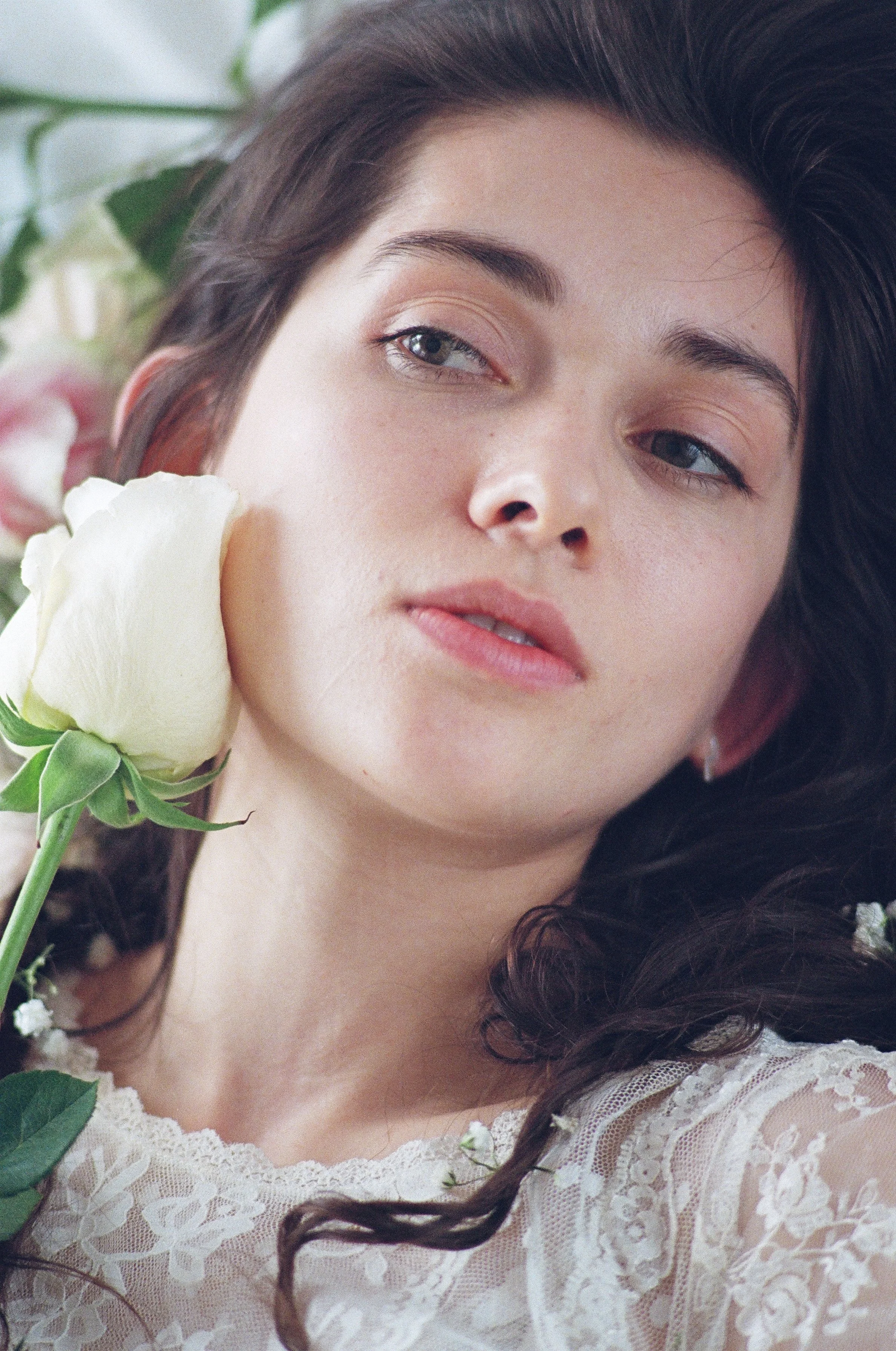 Close-up of a woman with dark hair, lying among white flowers, wearing a lace garment.