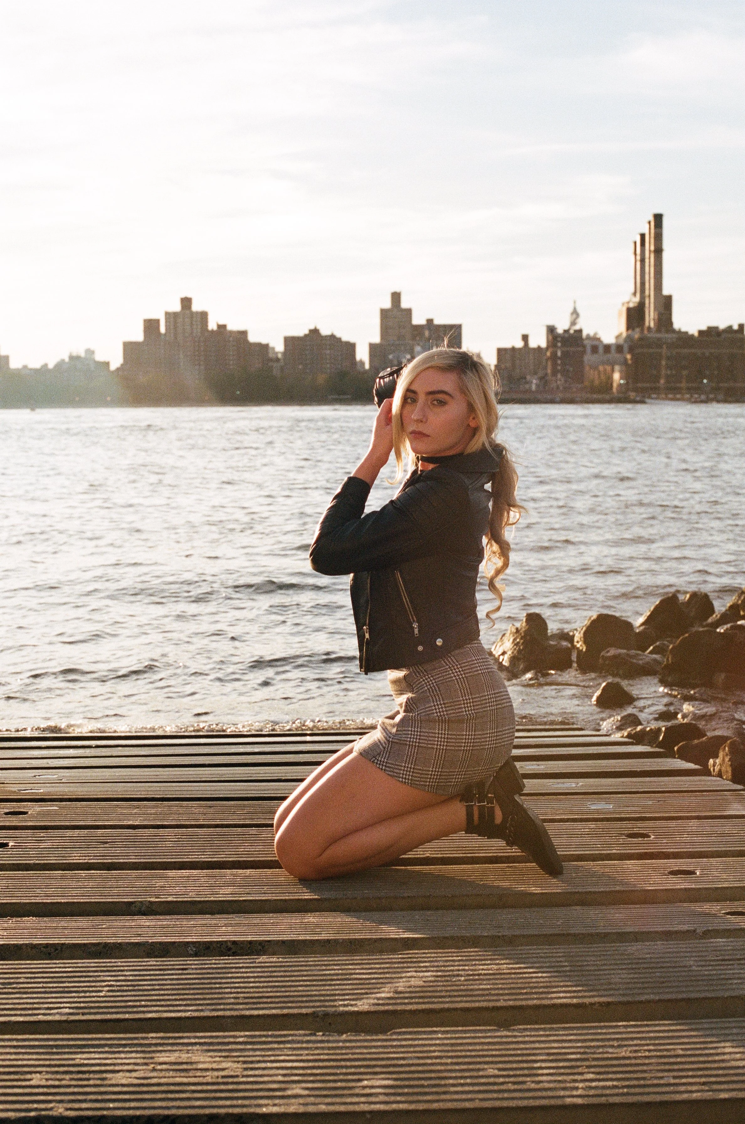 A young woman kneeling on a wooden dock by the water during sunset, with a city skyline in the background.